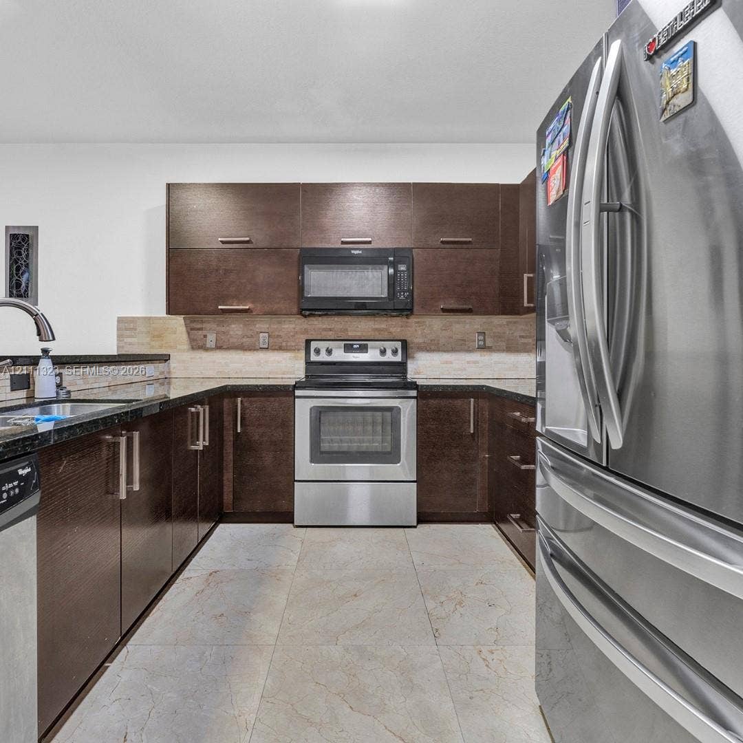 Kitchen featuring dark wood-finish cabinetry, stainless steel appliances, dark stone countertops, tile backsplash, and light-toned tile flooring