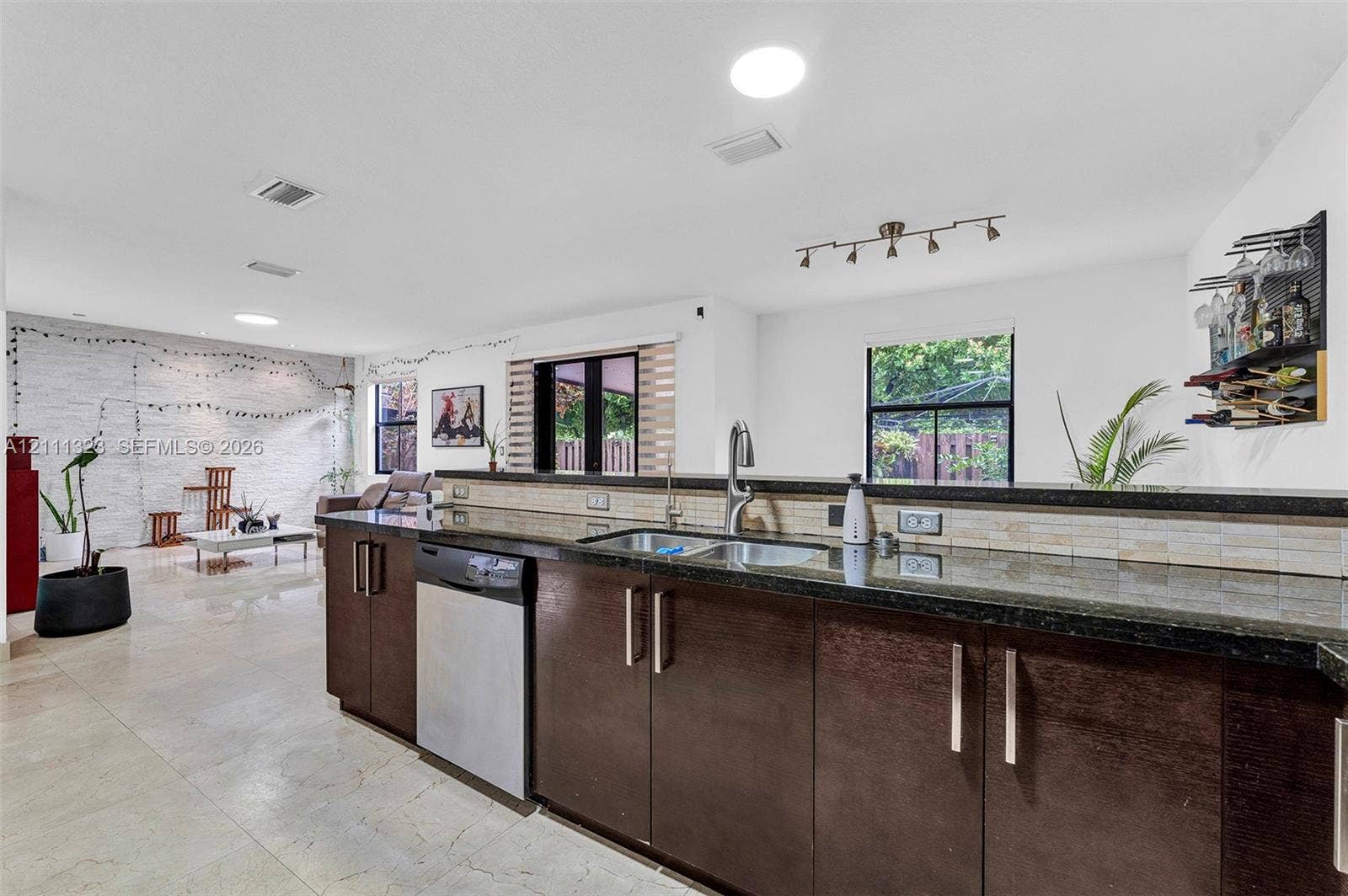 Kitchen featuring dark-toned flat-panel cabinetry, stone-finish countertops, stainless steel dual-basin sink with arched faucet, stainless steel dishwasher, and neutral tiled flooring