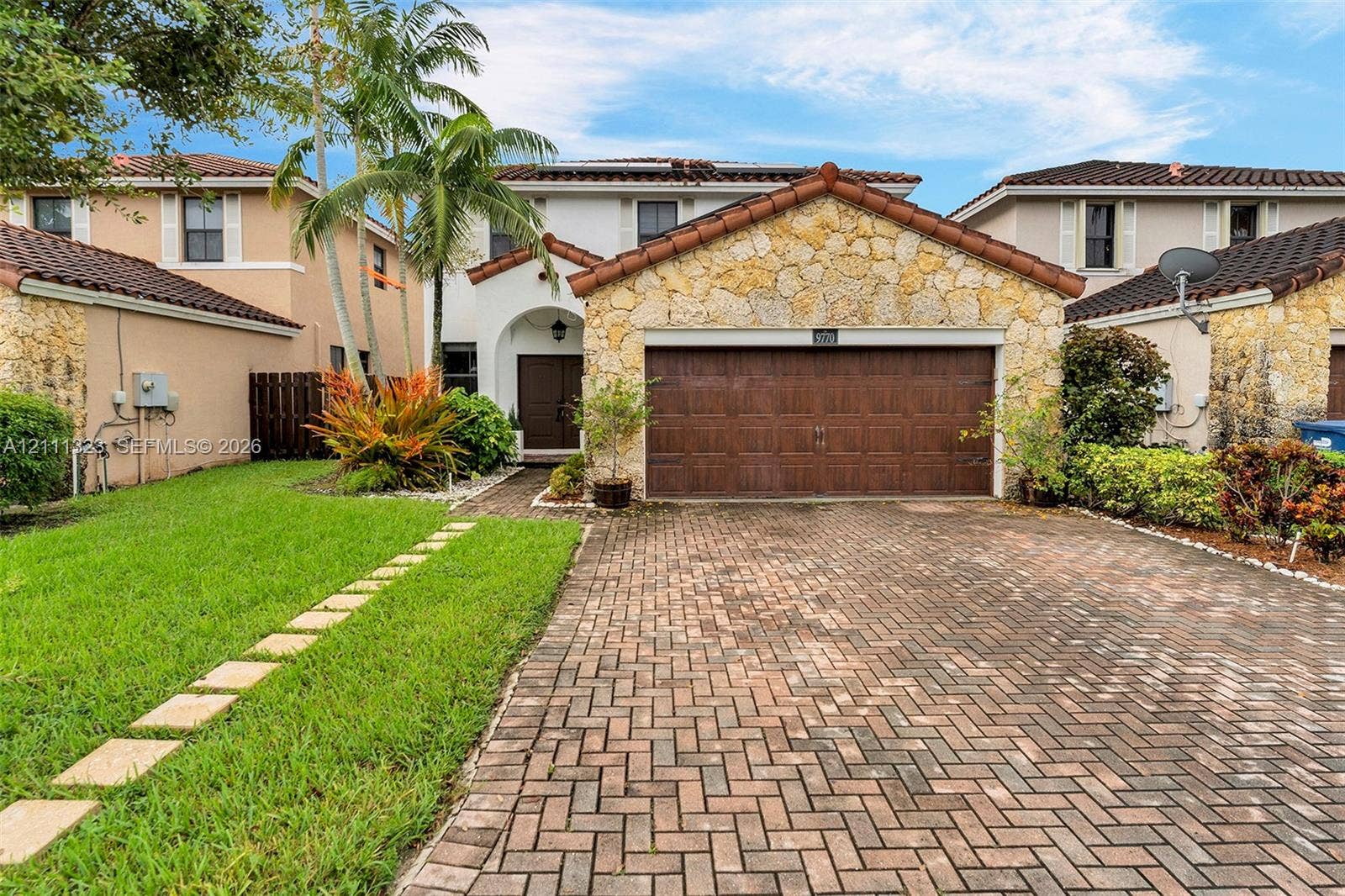 Two-story exterior featuring a natural stone facade, brown paneled garage door, arched entryway, brick-paver driveway, and manicured landscaping