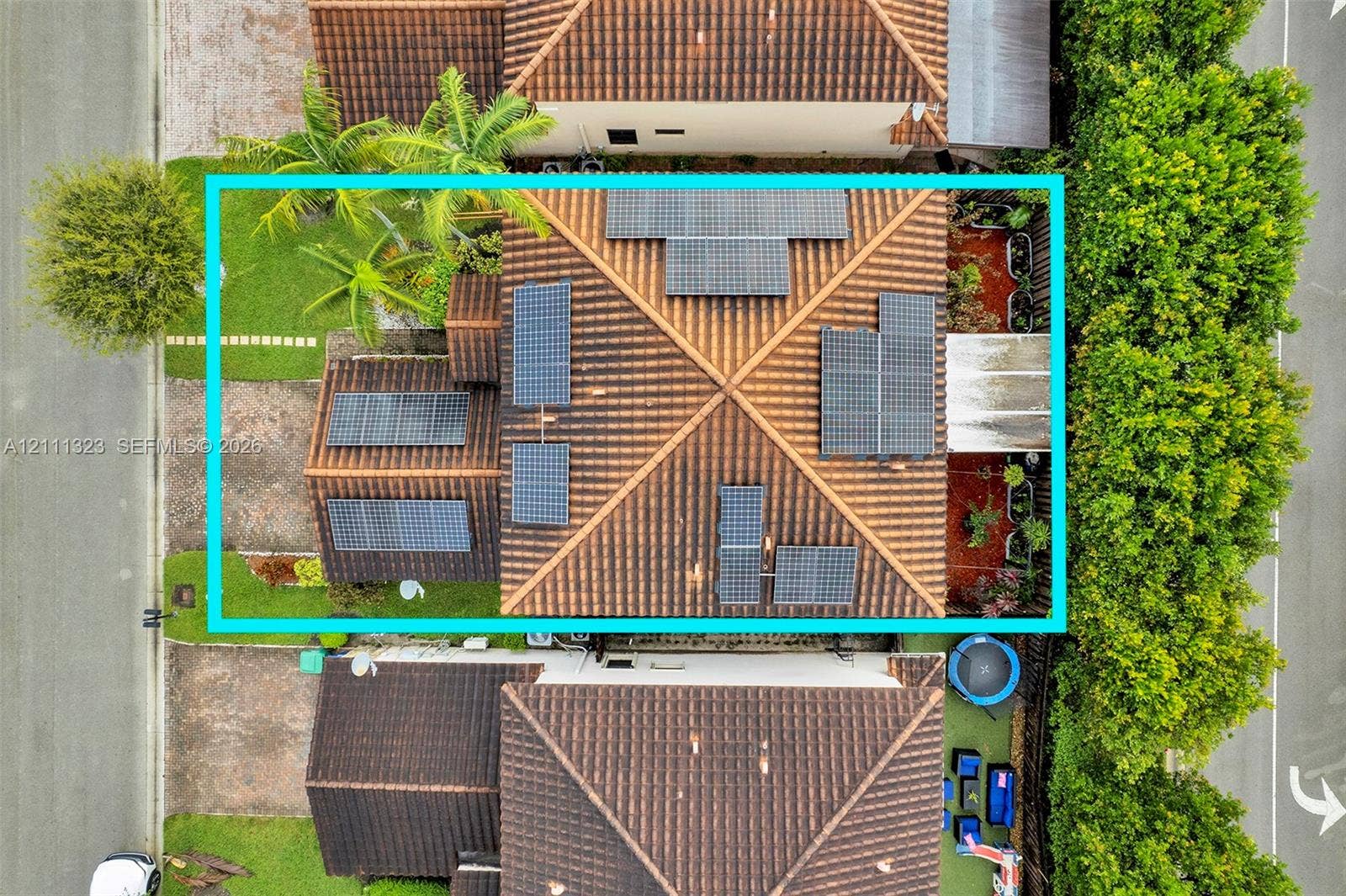 Aerial view of property featuring a tile roof equipped with a solar panel array, manicured landscaping, and mature palm trees