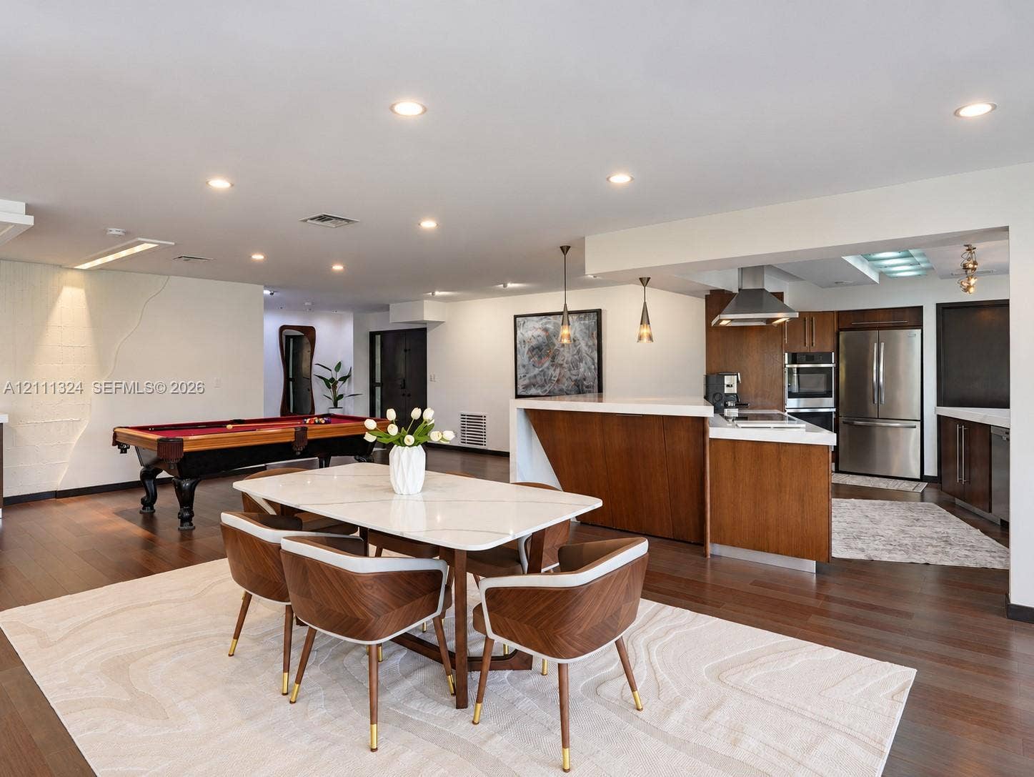 Open-concept kitchen featuring flat-panel wood cabinetry, integrated stainless steel appliances, a stainless steel range hood, and a kitchen island with a stone countertop, all illuminated by recessed lighting
