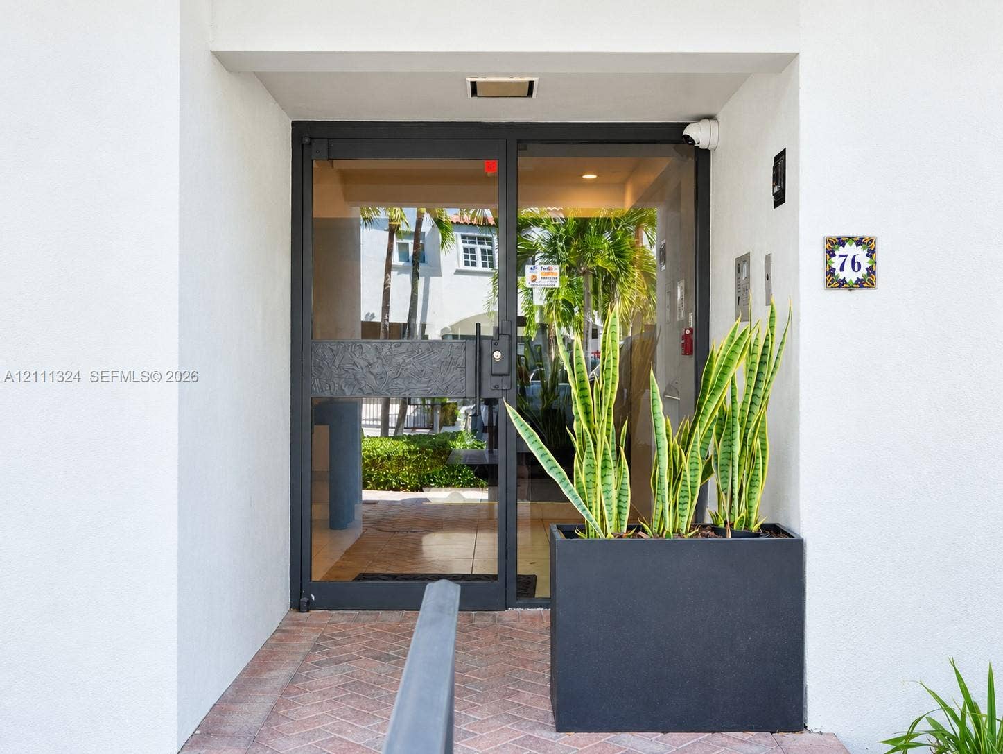 Entrance featuring a black metal-framed glass door, brick-paved walkway, recessed ceiling lighting, and a square address tile