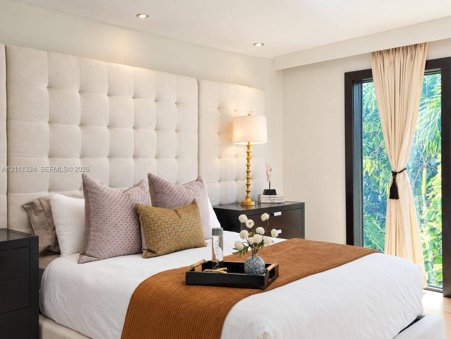 Tufted floor-to-ceiling fabric headboard, recessed ceiling lighting, and floor-to-ceiling window with dark-frame trim
