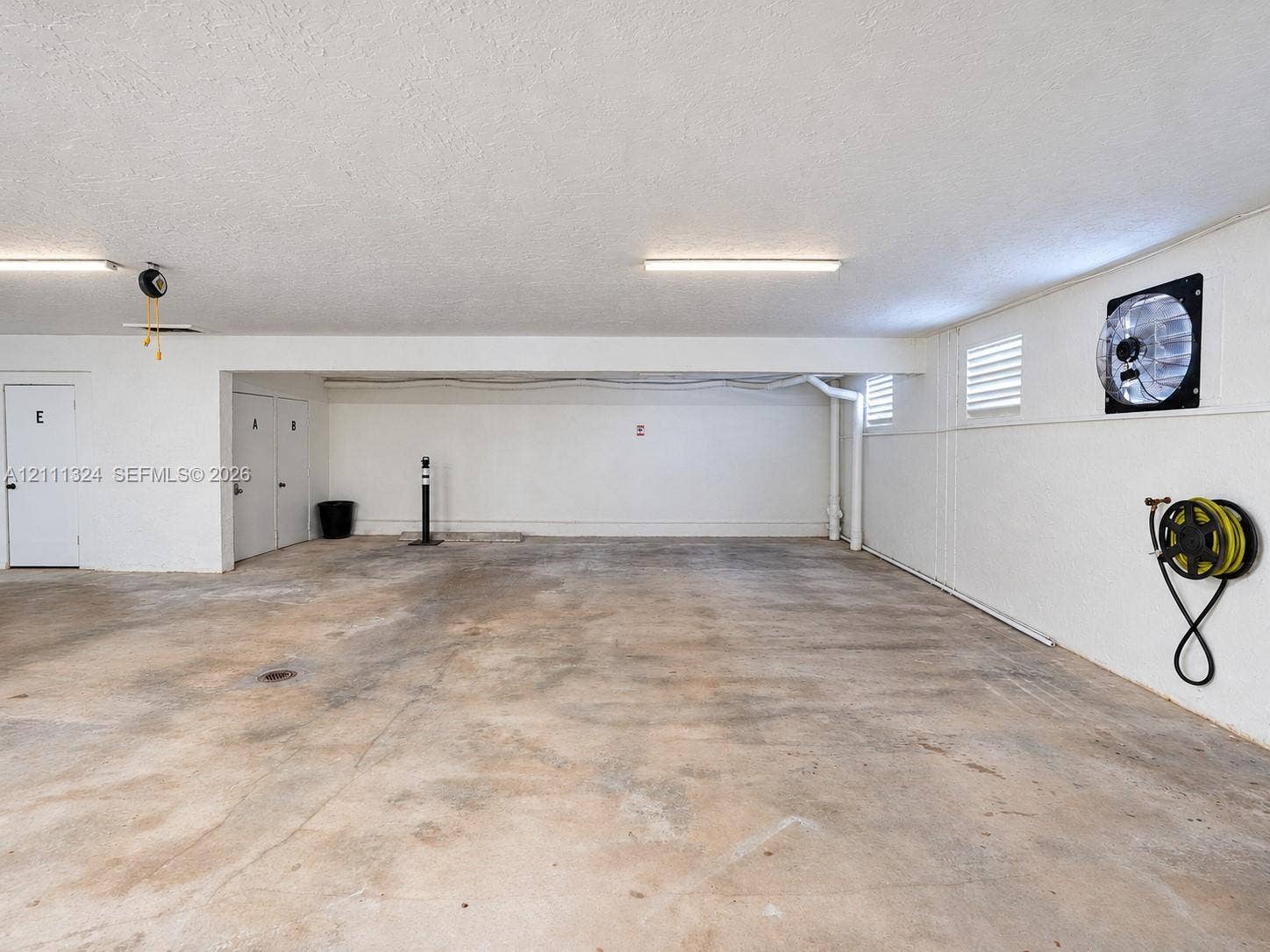 Covered parking area featuring concrete flooring, flush-mount lighting, louvered wall vents, and a mounted utility hose reel