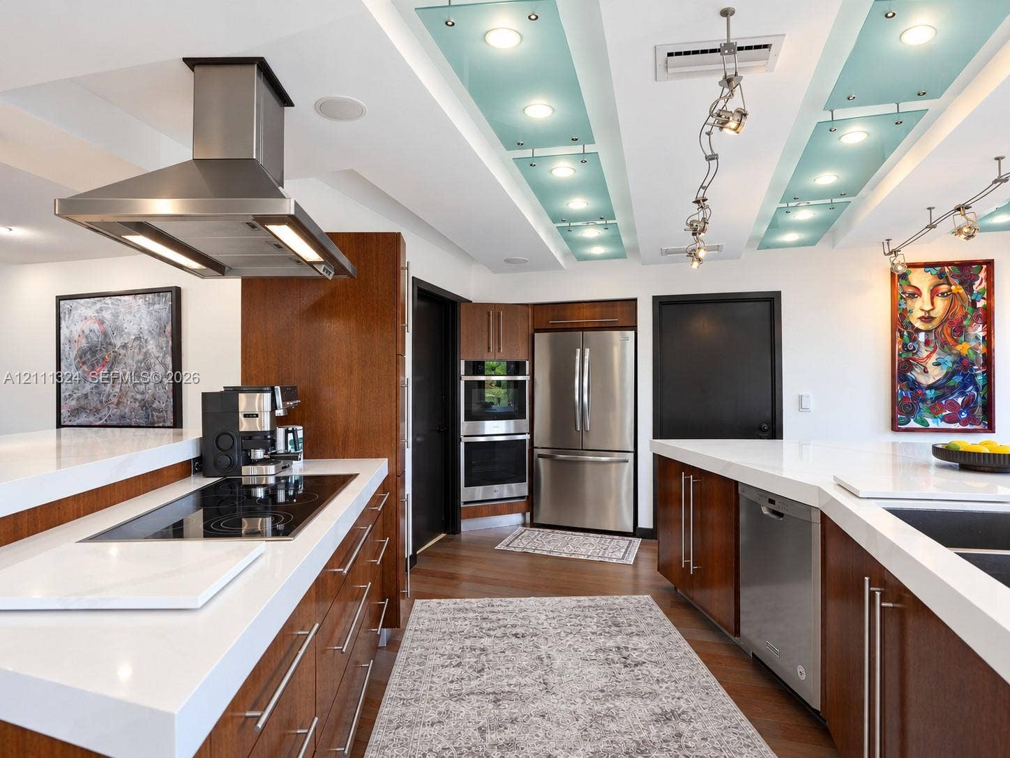 Kitchen featuring custom wood cabinetry, stainless steel appliances including a built-in double oven and range hood, white solid-surface countertops, and recessed lighting with decorative glass panels