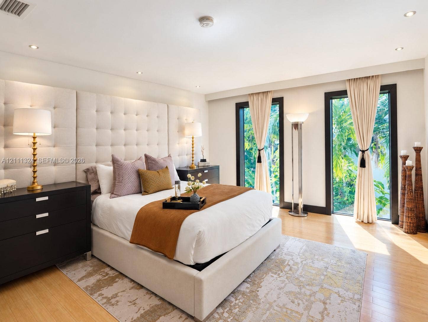 Floor-to-ceiling tufted fabric headboard, recessed ceiling lighting, and two full-length glass doors with dark frames and integrated drapery