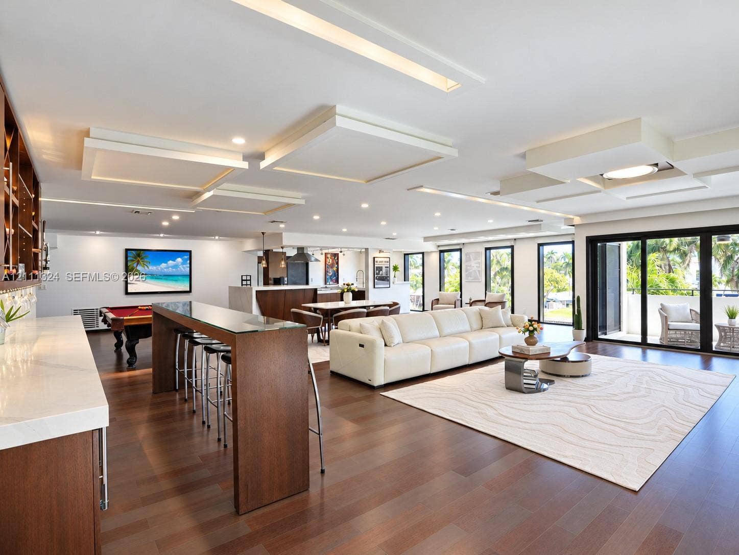 Open-concept living space featuring wood-finish flooring, custom coffered ceiling with recessed lighting, built-in bar with stone countertop, and floor-to-ceiling sliding glass doors