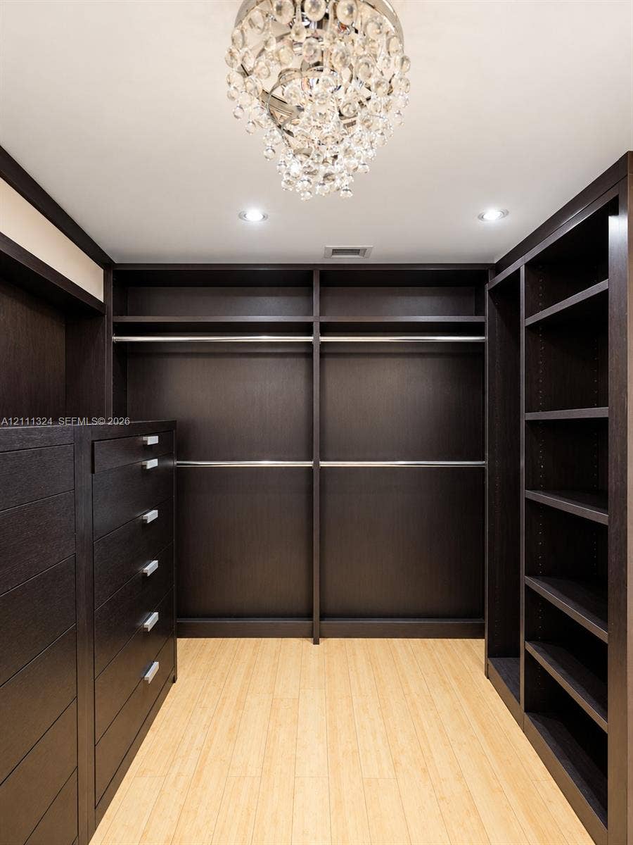 Custom walk-in closet featuring dark wood-finish built-in cabinetry, floor-to-ceiling shelving, integrated hanging rods, a central dresser unit with metallic pulls, and a crystal drop chandelier