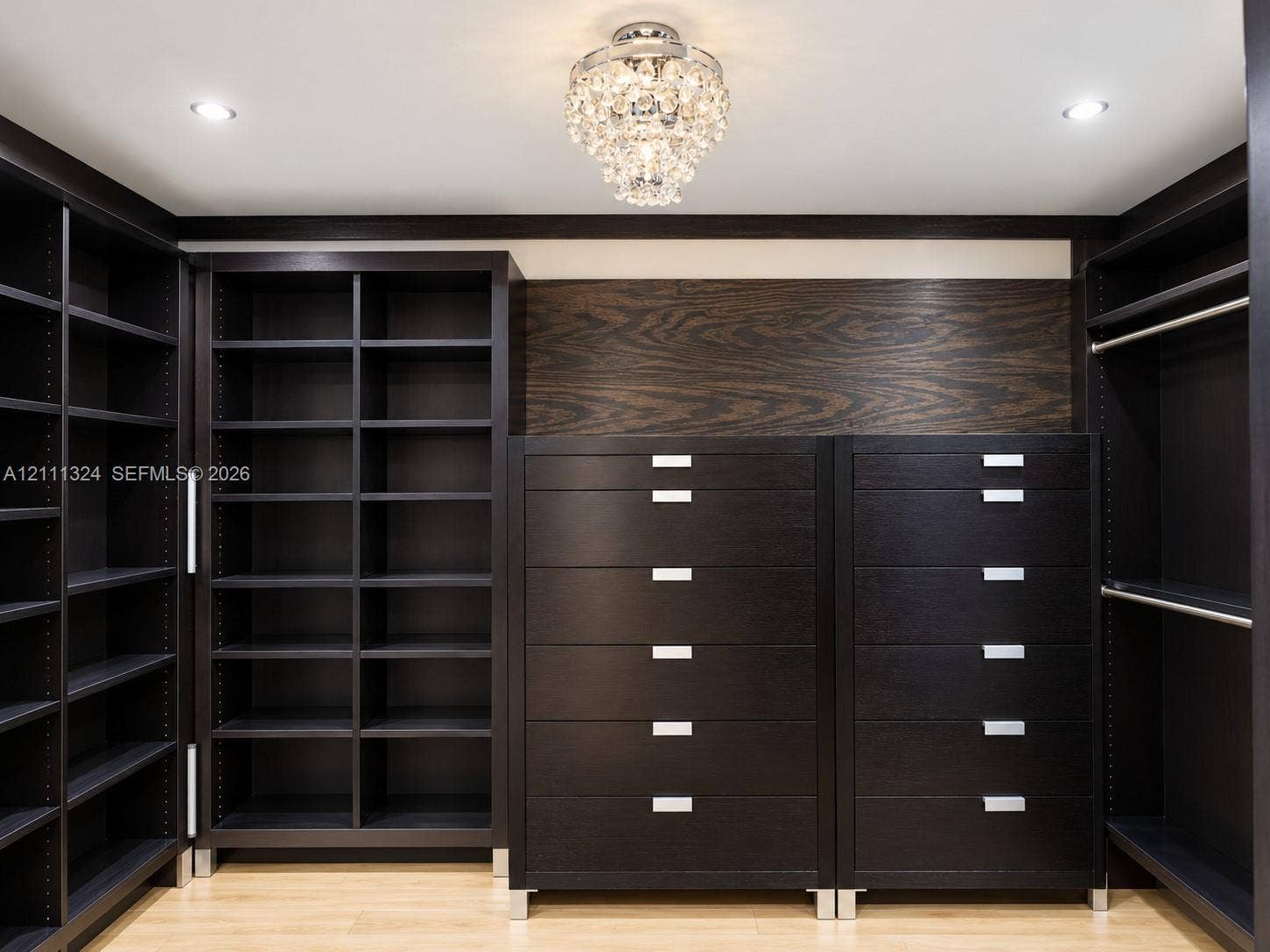Custom dark wood-finish built-in cabinetry featuring extensive shelving, integrated drawer units with metallic hardware, and a center decorative crystal chandelier