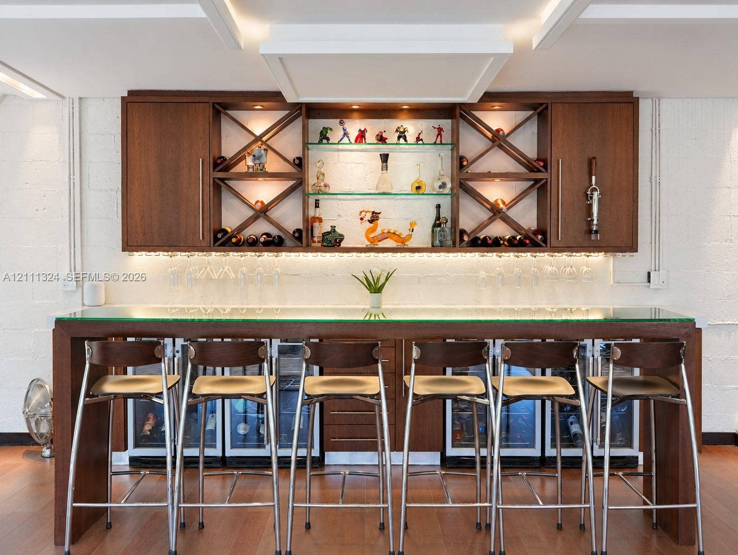 Custom built-in bar featuring wood cabinetry, integrated glass shelving with X-frame detailing, under-cabinet lighting, and a glass-top counter with multiple under-counter refrigeration units