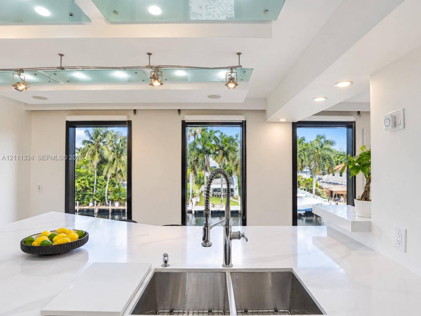Kitchen featuring a white stone-finish countertop, dual-basin stainless steel sink with a high-arch gooseneck faucet, ceiling-mounted track lighting with glass panels, and windows displaying water and palm tree scenery