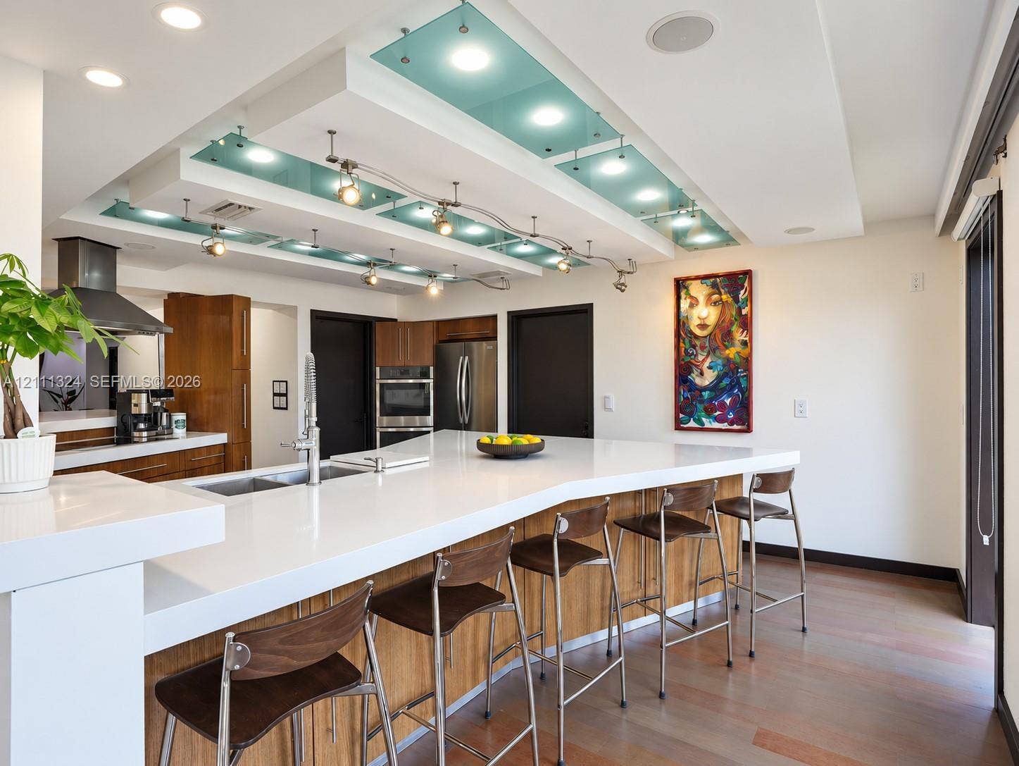 Kitchen featuring a custom tiered drop ceiling with integrated glass panels, recessed lighting, and adjustable track lighting, paired with an expansive island with a stone-surface countertop, built-in dual stainless steel ovens, and wood-finish cabinetry