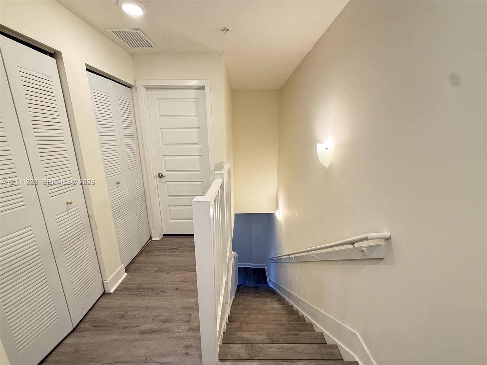 10443 NW 82nd Street, #6, Doral, Doral, FL 33178 Hallway featuring wood-finish flooring, two sets of louvered bifold closet doors, a multi-panel interior door, and wall-mounted sconce lighting