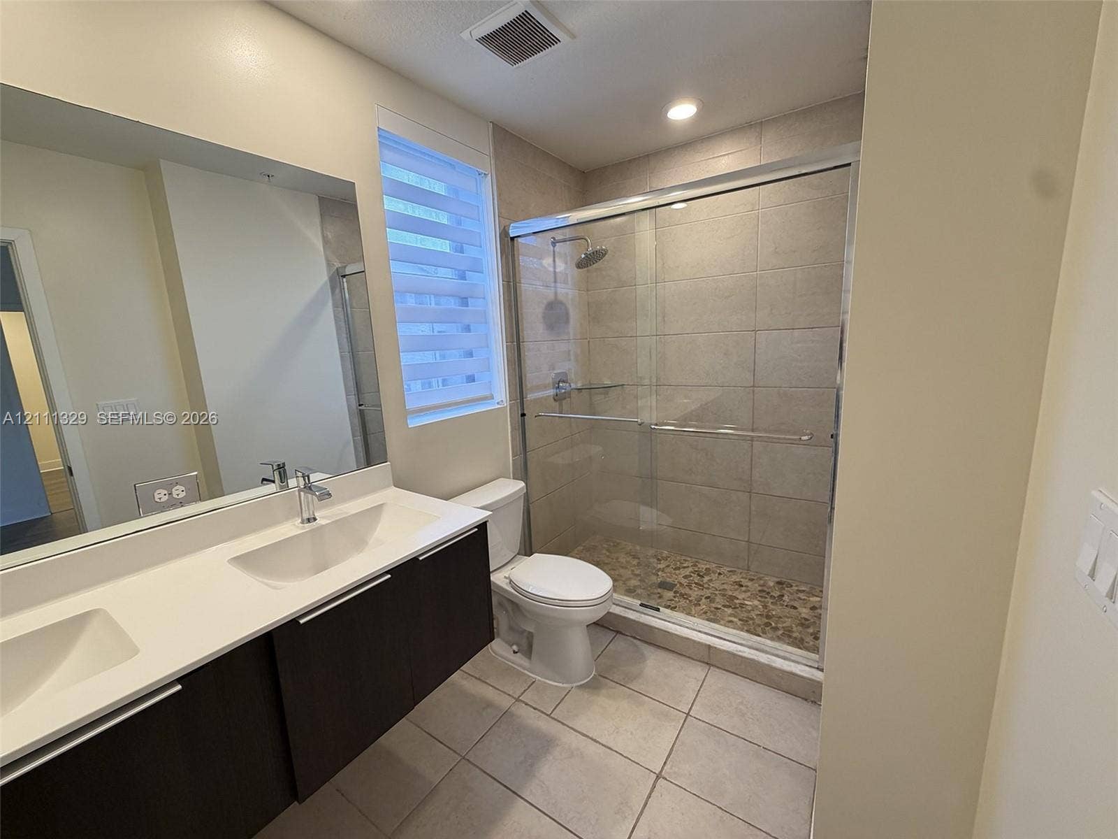 10443 NW 82nd Street, #6, Doral, Doral, FL 33178 Double vanity featuring a light-toned countertop with integrated rectangular basins, dark wood-finish cabinetry, a large frameless mirror, and a walk-in shower with stone tile flooring and glass sliding doors