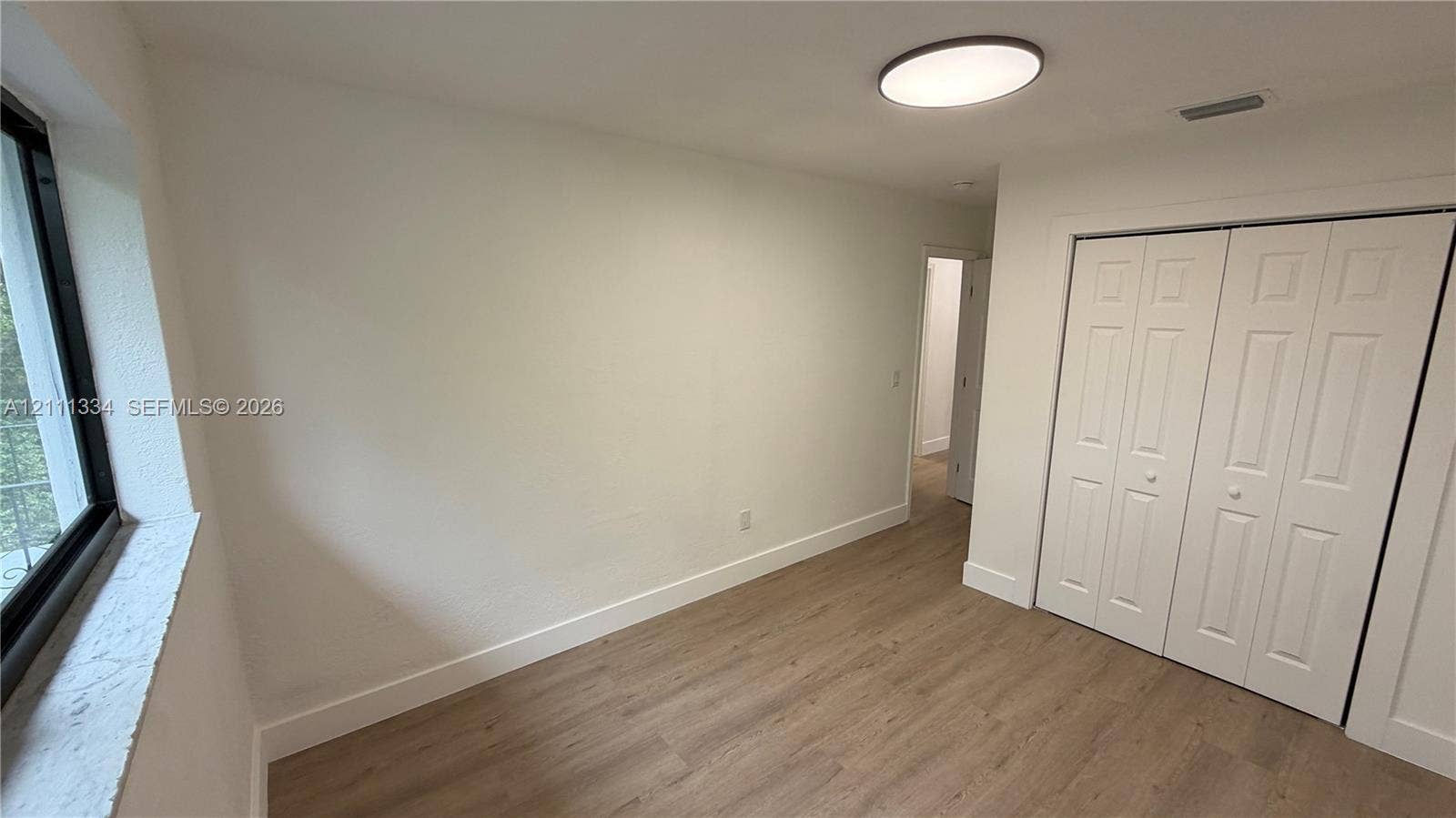 3314 NW 8th, #1, Miami, Miami, FL 33131 Interior space featuring wood-finish flooring, a large window with a deep sill, neutral wall finishes, a flush-mount circular lighting fixture, and a white bi-fold closet door