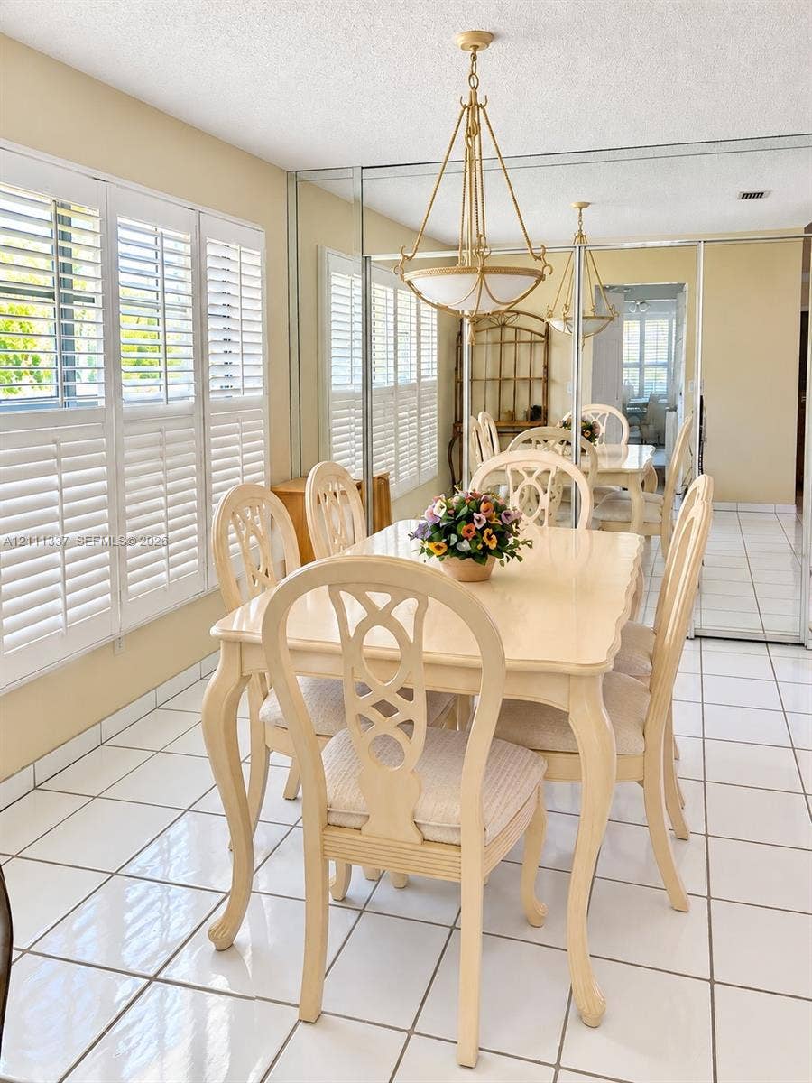 Dining space featuring white ceramic tile flooring, full-wall mirrored panels, plantation shutters, and a multi-arm metal chandelier