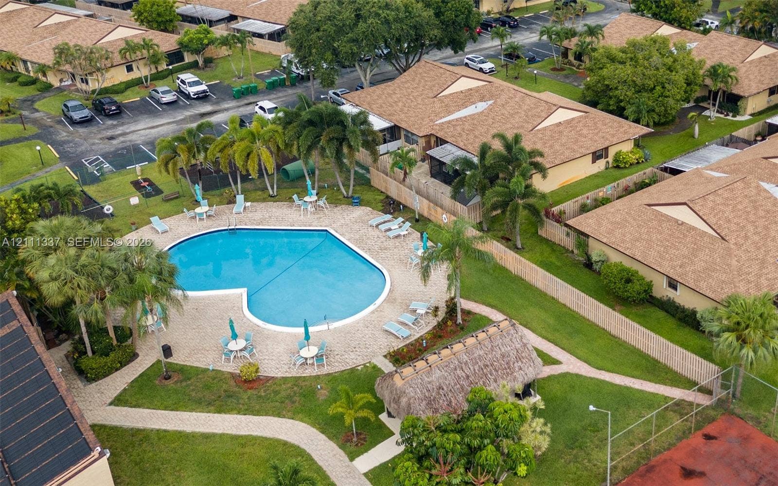 Community swimming pool featuring a paved deck, thatched-roof cabana, mature palm trees, and an enclosed tennis court