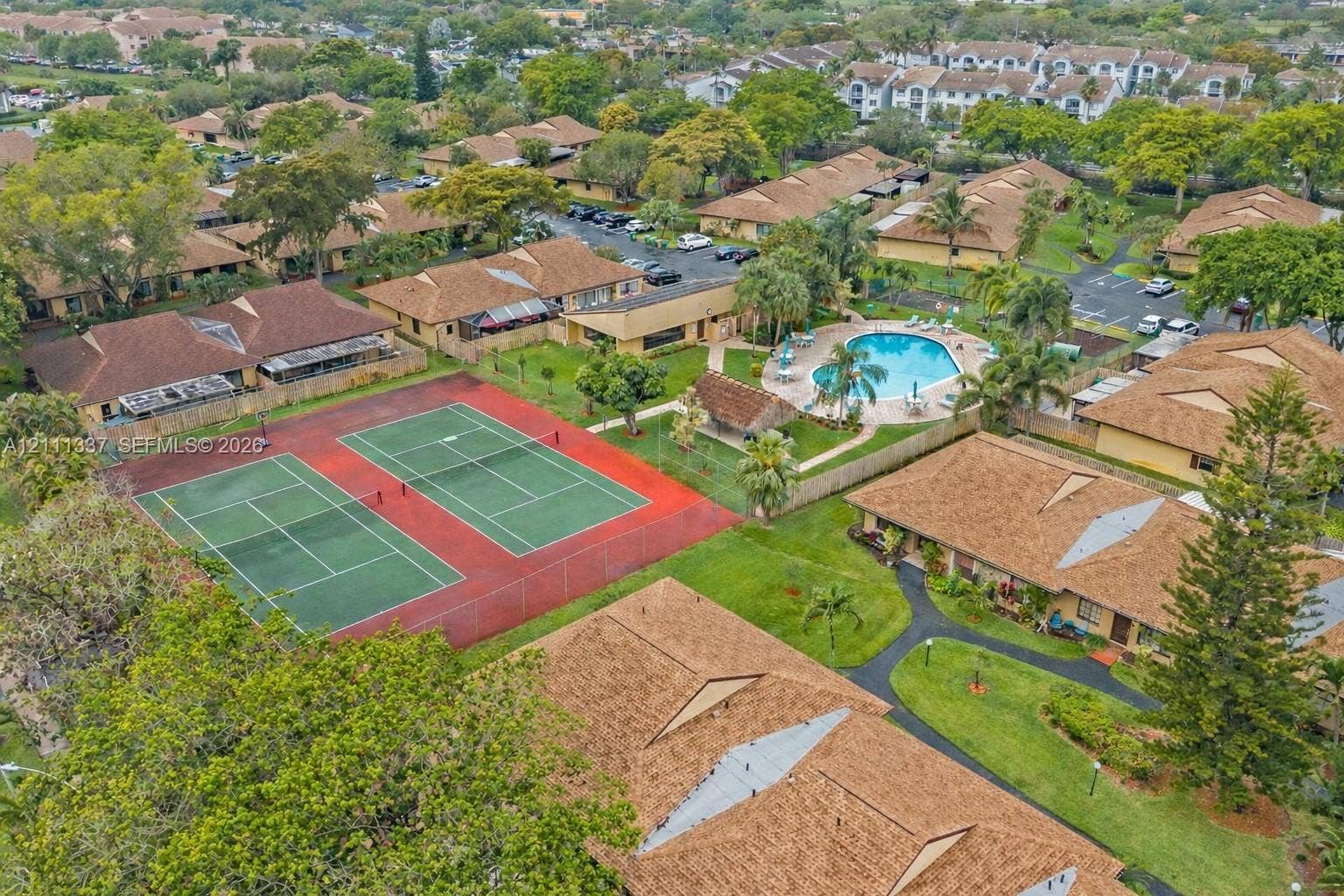 Aerial perspective of a residential community featuring dual outdoor tennis courts, a swimming pool, pedestrian walkways, and mature landscaping