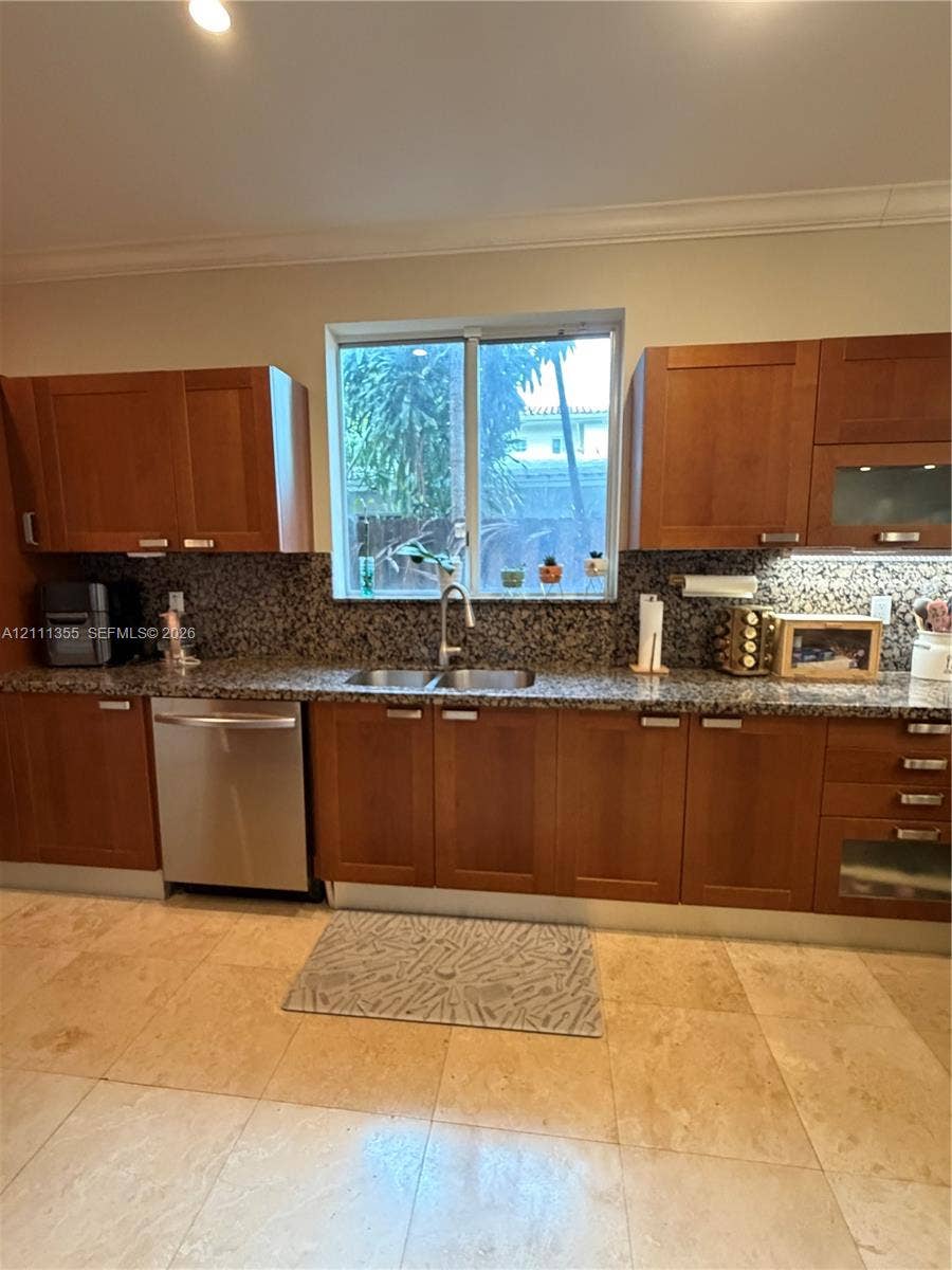Kitchen featuring warm-toned wood cabinetry, natural stone countertops, a full-height stone backsplash, stainless steel dishwasher, and large-format tile flooring