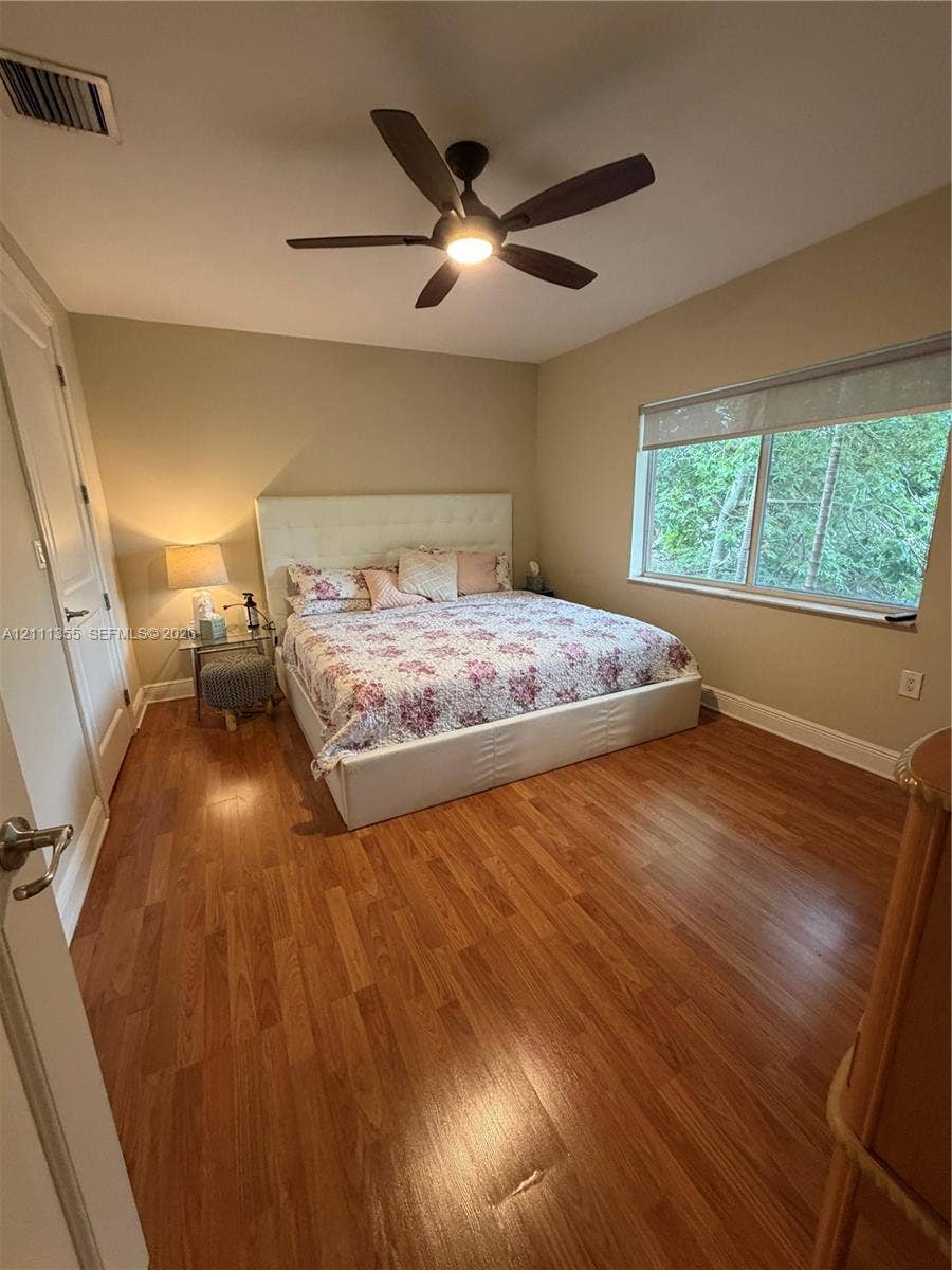 Room featuring wood-finish flooring, a multi-blade ceiling fan with integrated lighting, and a large window with a roller shade