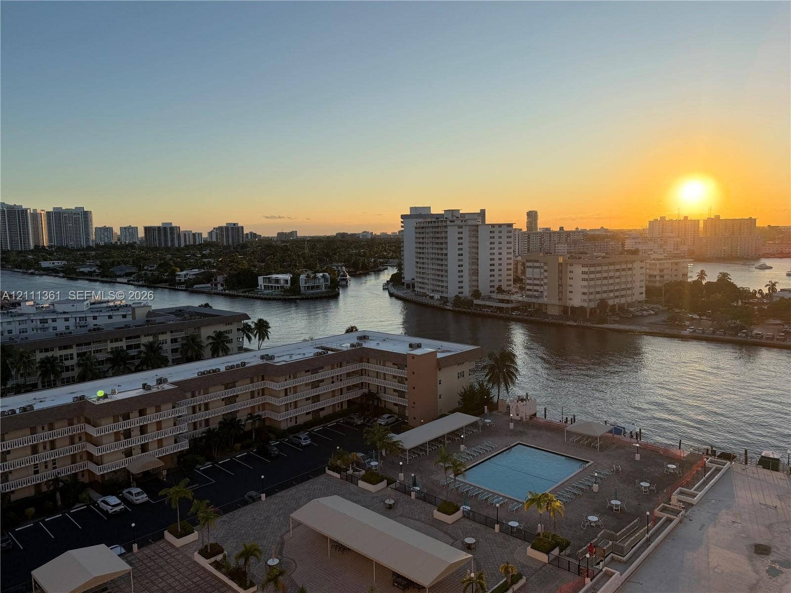 Elevated perspective overlooking a waterfront canal, swimming pool with deck space, covered parking areas, and a horizon featuring a coastal cityscape