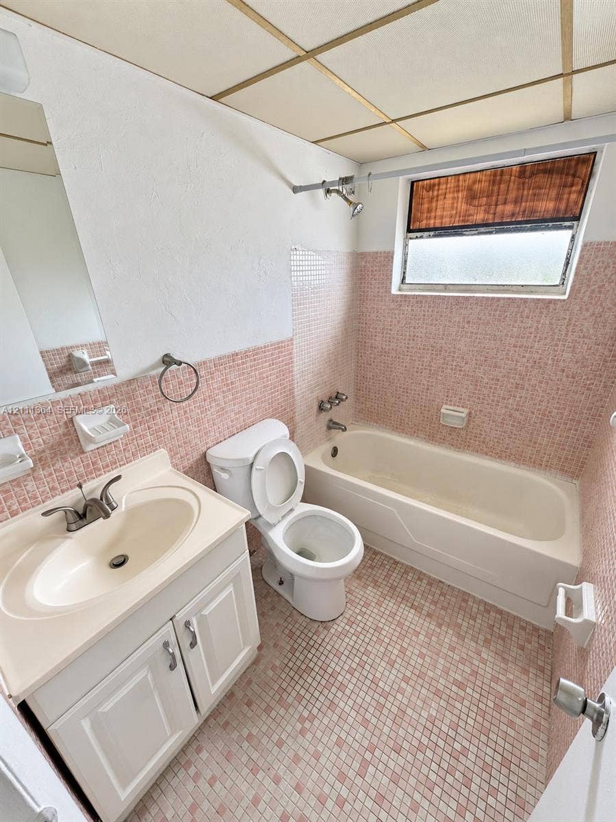 Bathroom featuring pink mosaic tile flooring and wall accents, a white vanity with an integrated basin, a standard bathtub with a shower head, and a drop ceiling with integrated lighting