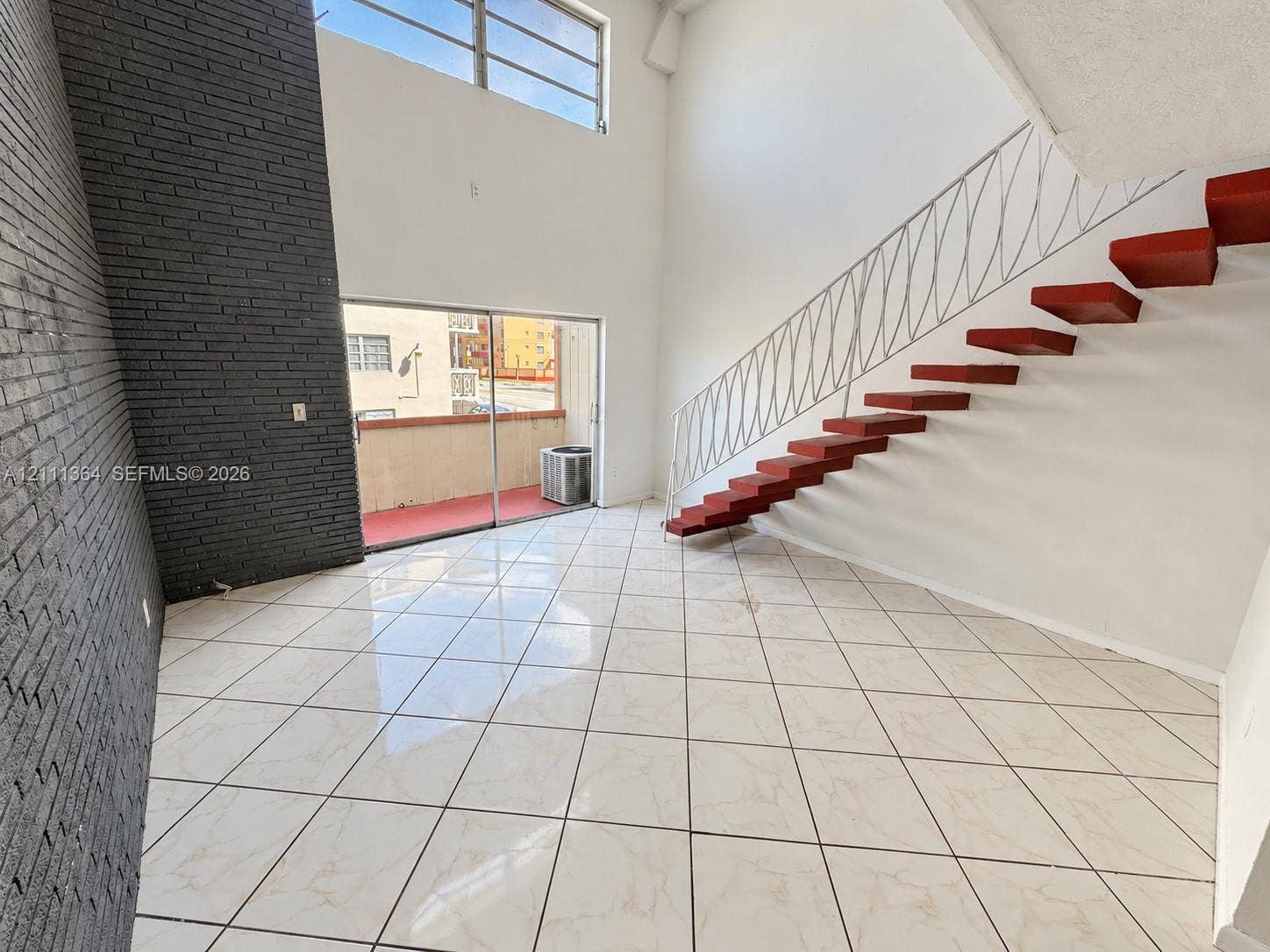Double-height ceiling, decorative dark-toned brick accent wall, ceramic tile flooring, floating staircase with custom iron railing, and sliding glass doors