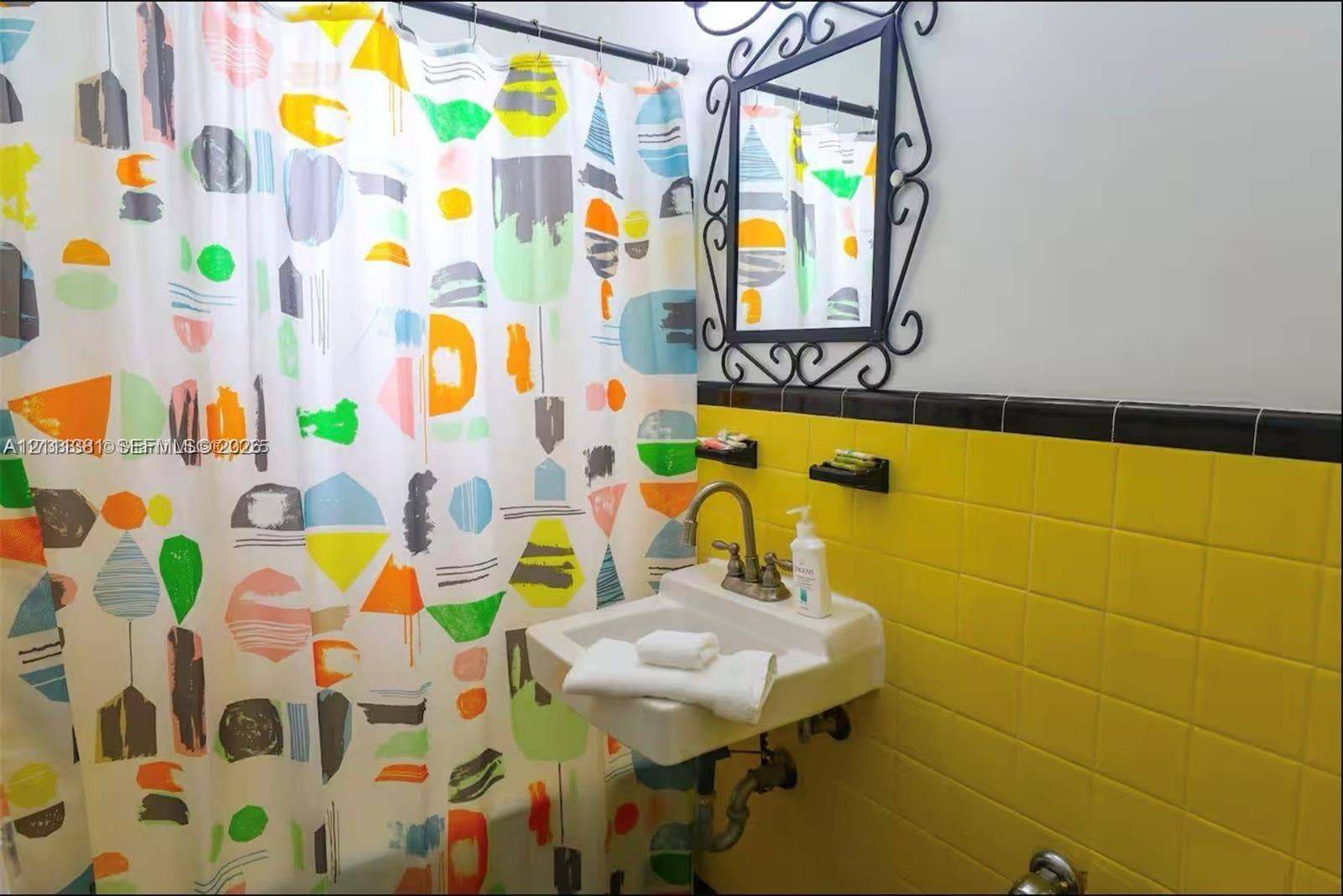 3710 Collins Avenue, #N-206, Miami Beach, FL 33140 Wall-mounted porcelain sink with dual-handle metal faucet, yellow ceramic tile wainscoting with black trim, and decorative wrought-iron framed wall mirror