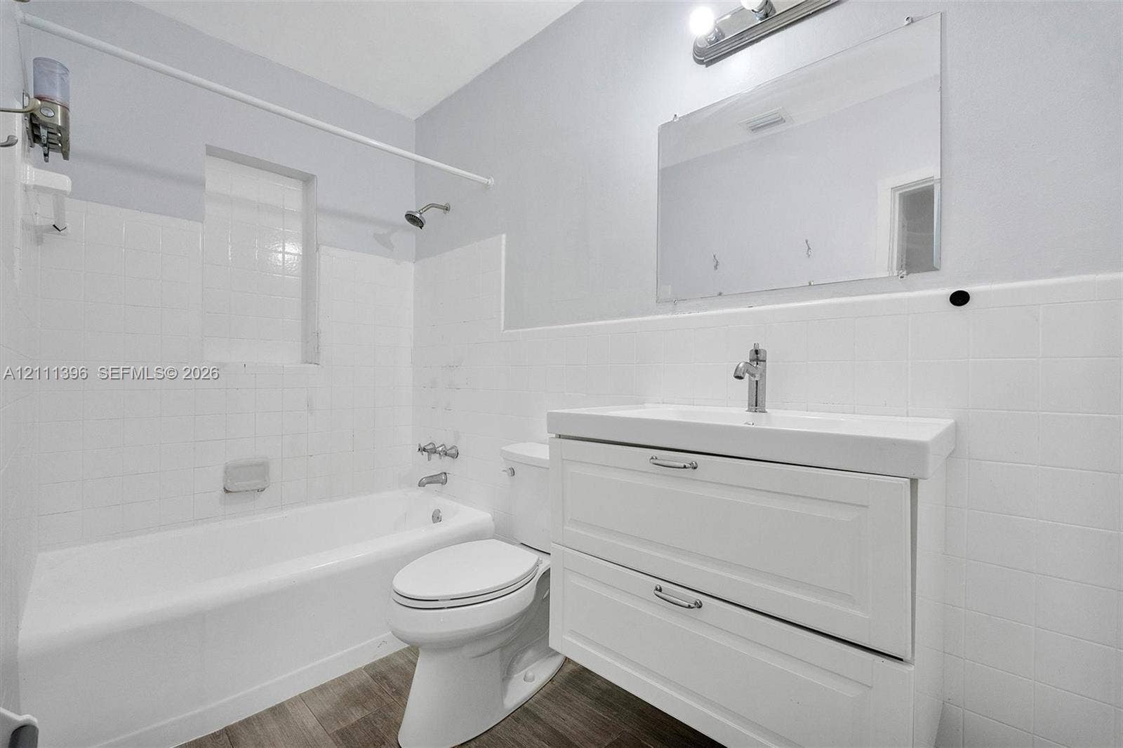 Bathroom featuring a white vanity with integrated basin, polished chrome faucet, frameless wall-mounted mirror, wood-finish flooring, and white tiled tub surround