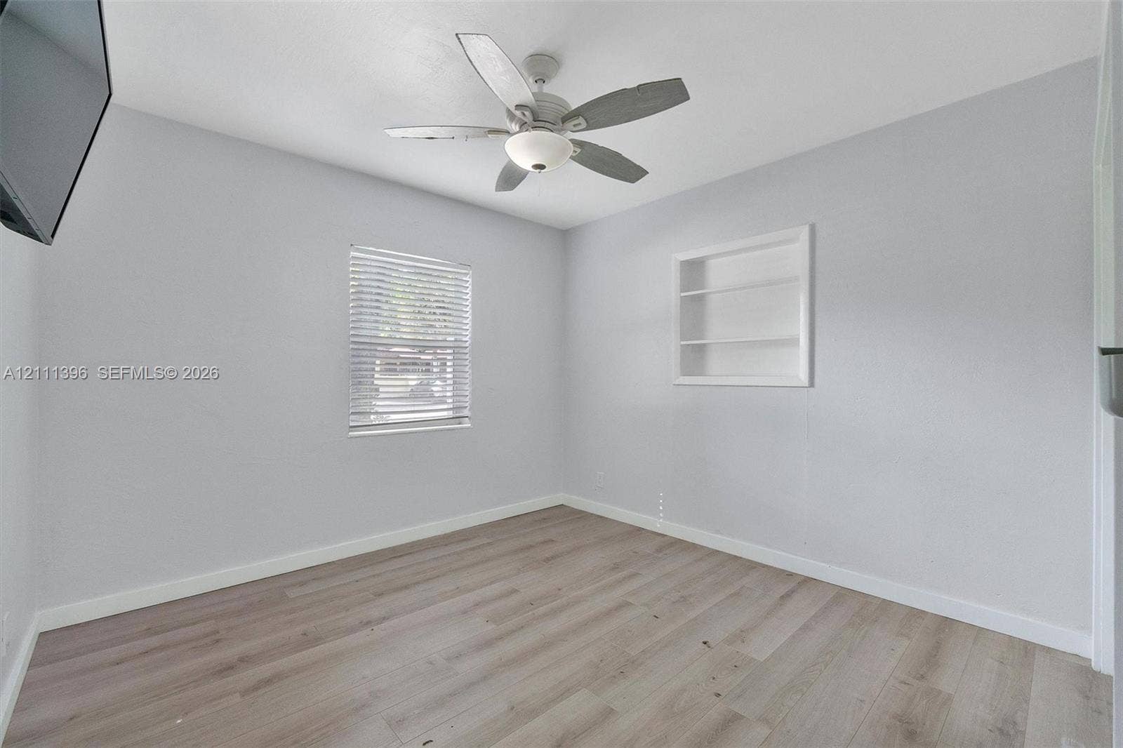 Room featuring wood-finish flooring, a ceiling fan with integrated light fixture, a window with horizontal blinds, and built-in wall shelving