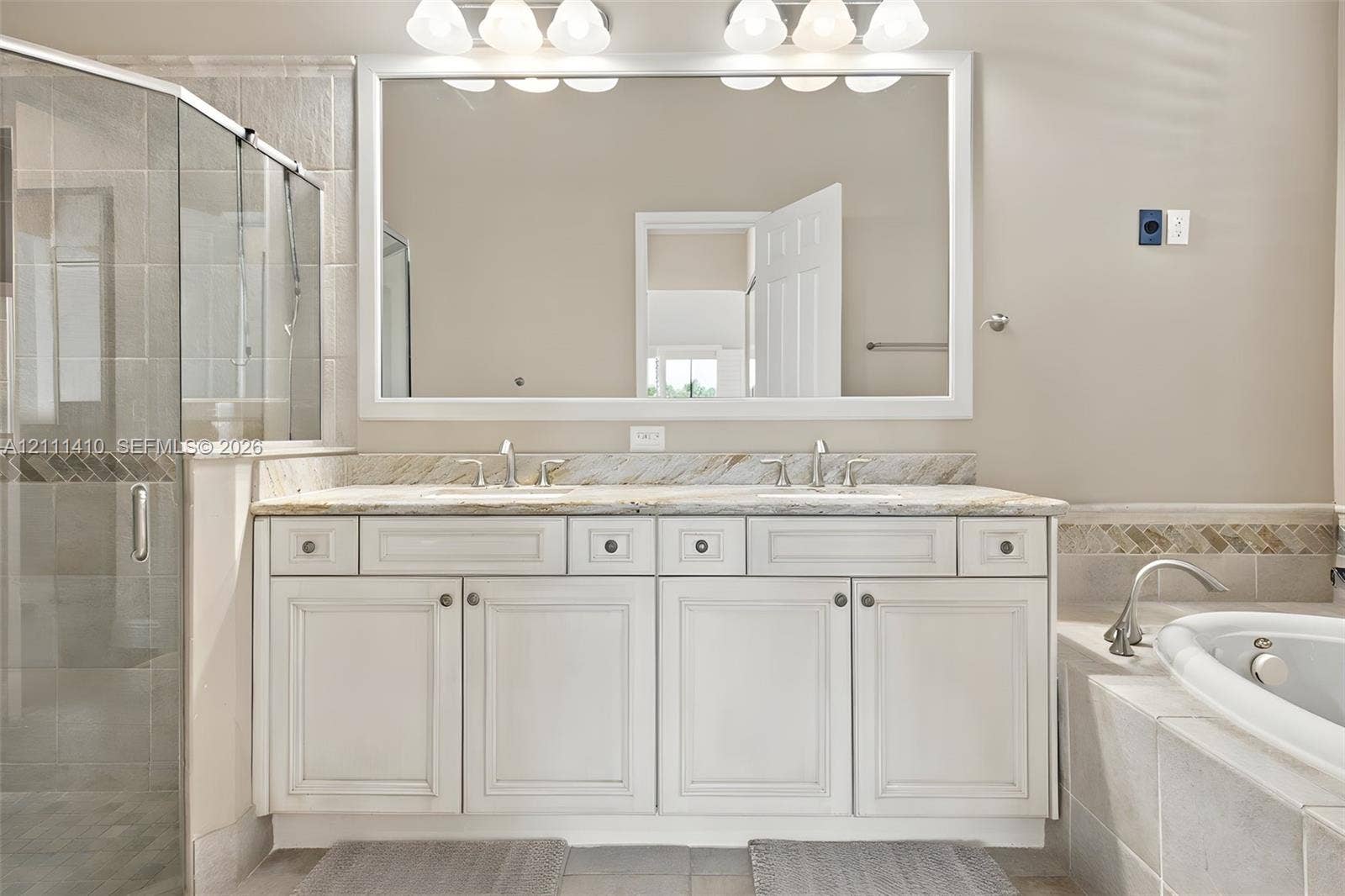 Dual-sink vanity with stone countertop, large framed wall mirror, glass-enclosed shower, and deep soaking tub with tile surround