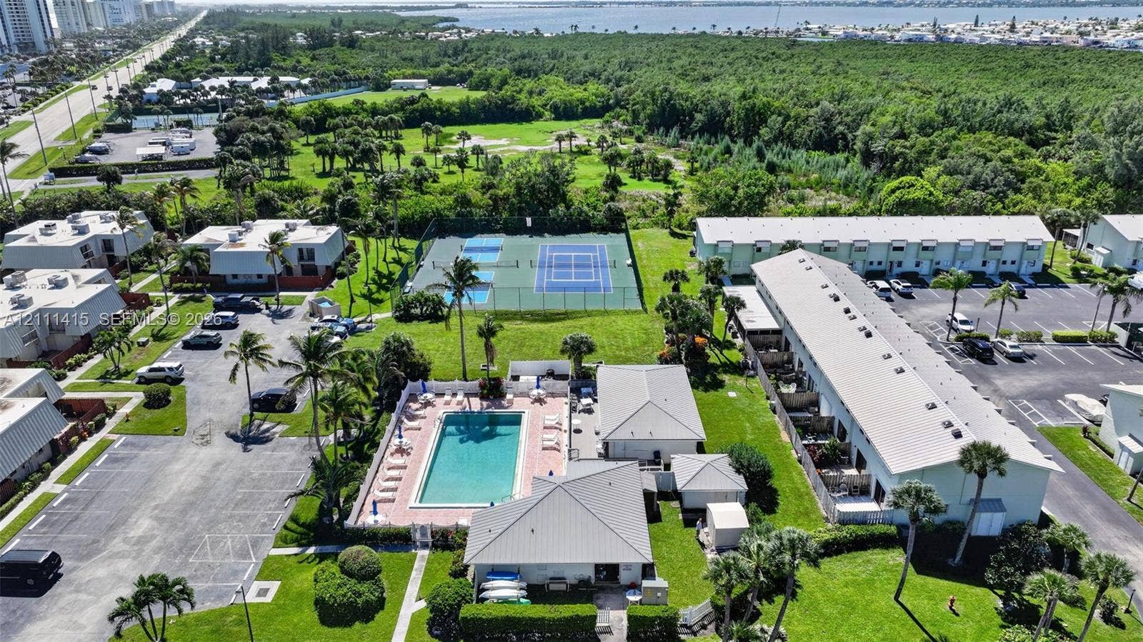 9423 S Ocean Drive, #80, Jensen Beach, FL 34957 Aerial perspective of multi-family complex featuring a swimming pool with paved deck, two fenced tennis courts, expansive green space, and mature palm trees