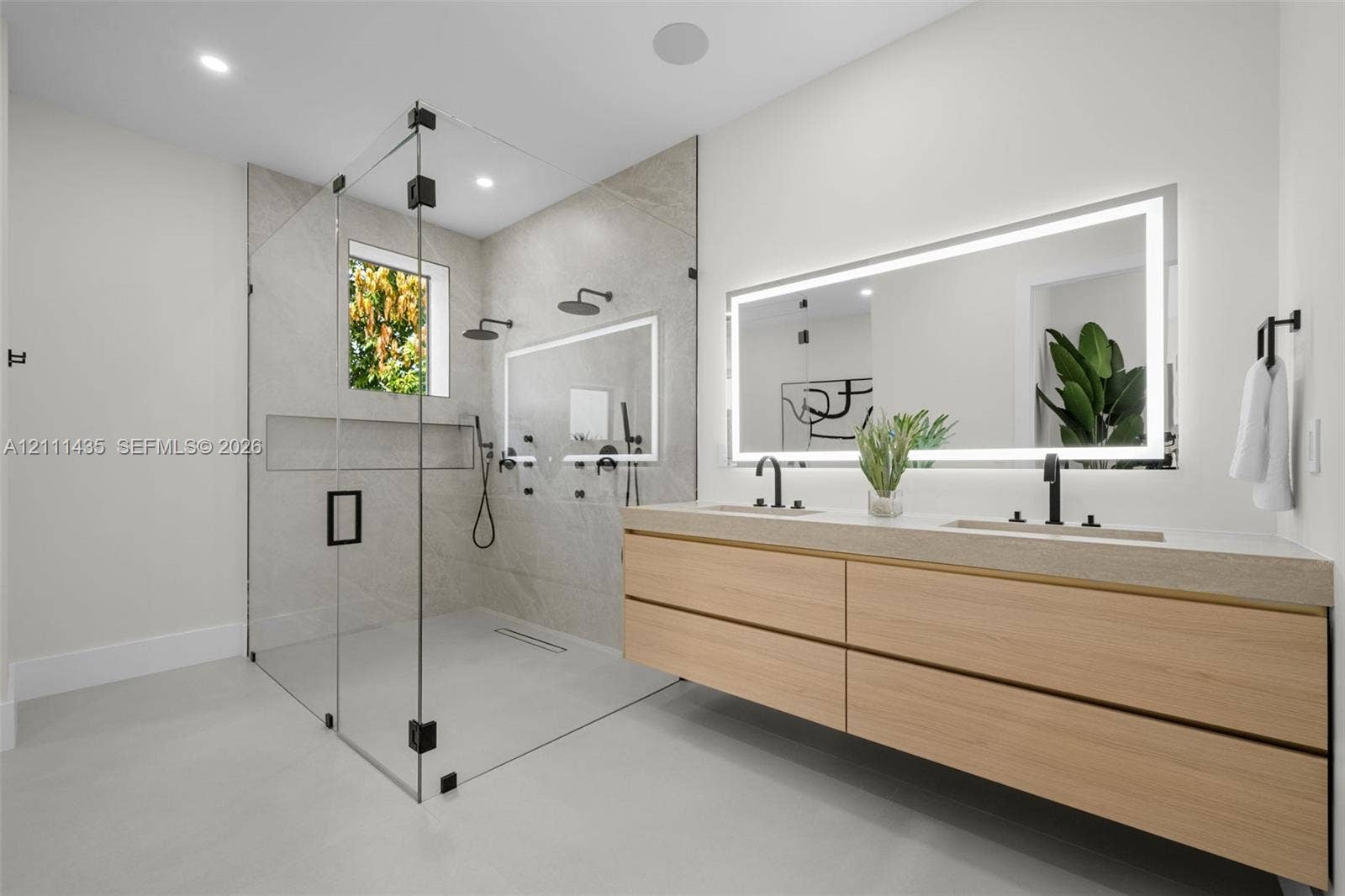 9530 SW 95th Court, Miami, FL 33176 Floating dual-sink vanity with light wood cabinetry and solid-surface countertop, frameless glass walk-in shower with stone tile surround, integrated recessed shower niche, and rectangular wall-mounted backlit mirror