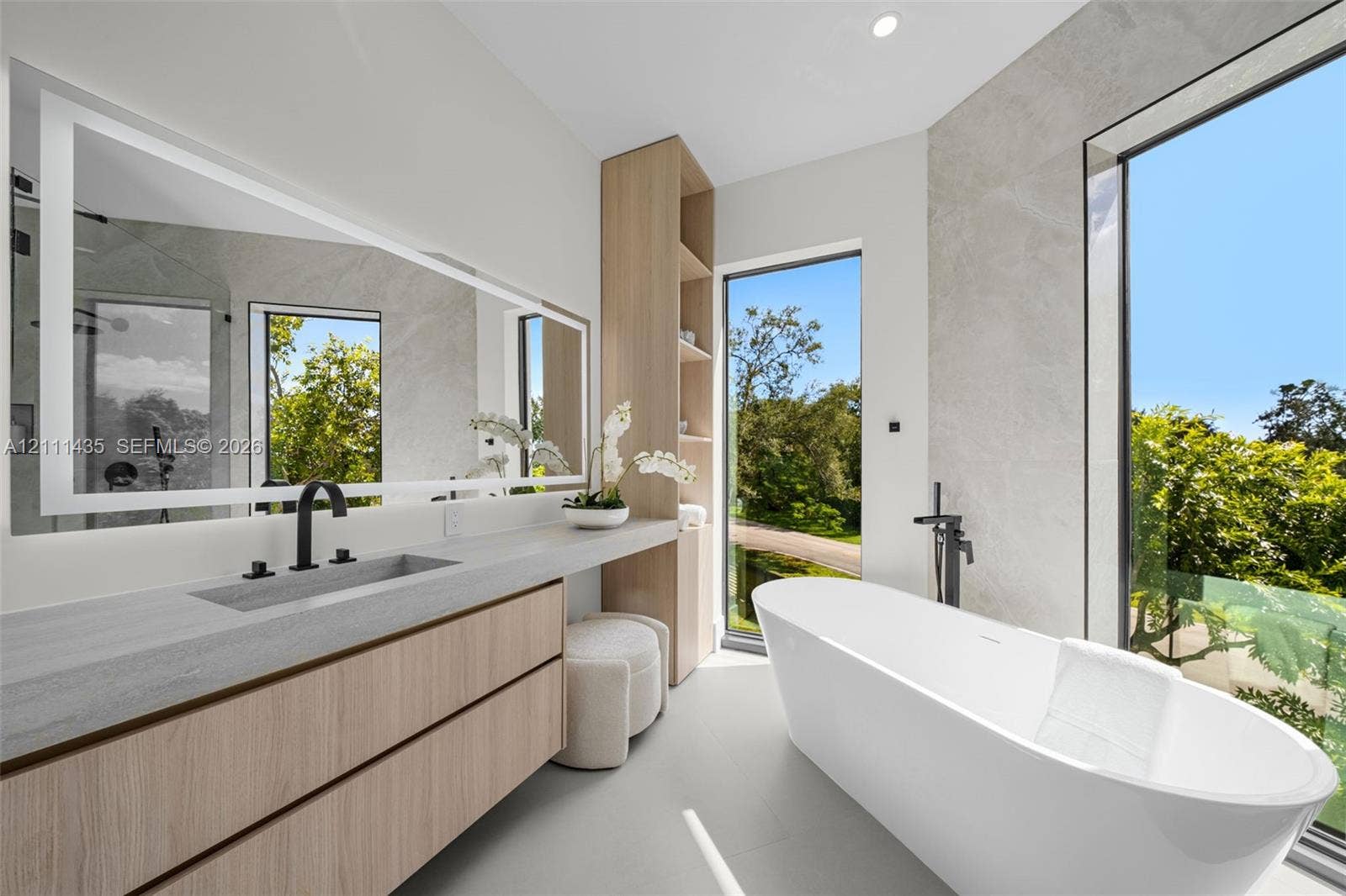 9530 SW 95th Court, Miami, FL 33176 Floating wood-finish vanity with integrated sink, expansive LED-backlit mirror, freestanding soaking tub with floor-mounted faucet, and floor-to-ceiling windows
