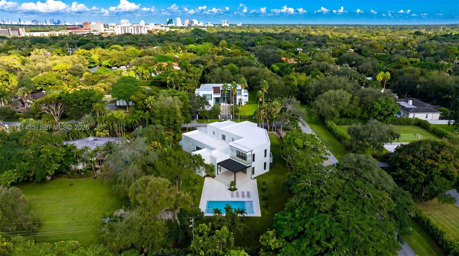 9530 SW 95th Court, Miami, FL 33176 Multi-level modern residence featuring a white exterior, a covered rear patio, a swimming pool, and a manicured lawn set within a wooded lot