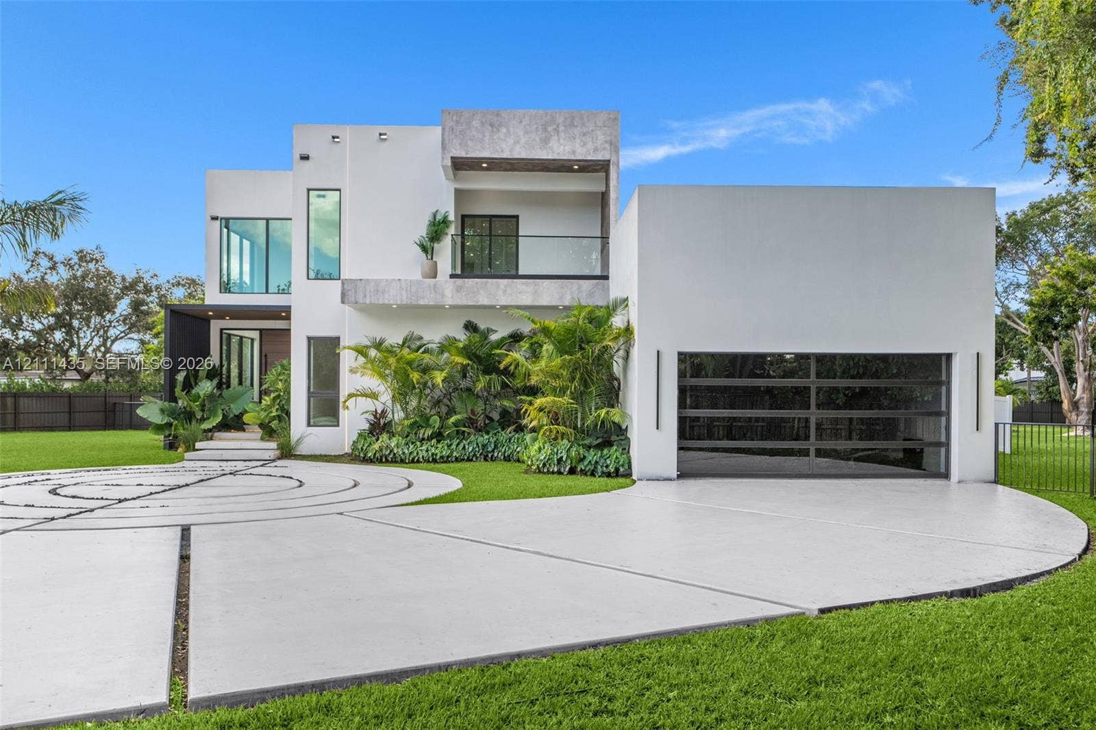9530 SW 95th Court, Miami, FL 33176 Modern two-story exterior featuring smooth stucco siding, a second-level balcony with glass railing, tiered concrete driveway with decorative inlay, and an oversized multi-panel garage door