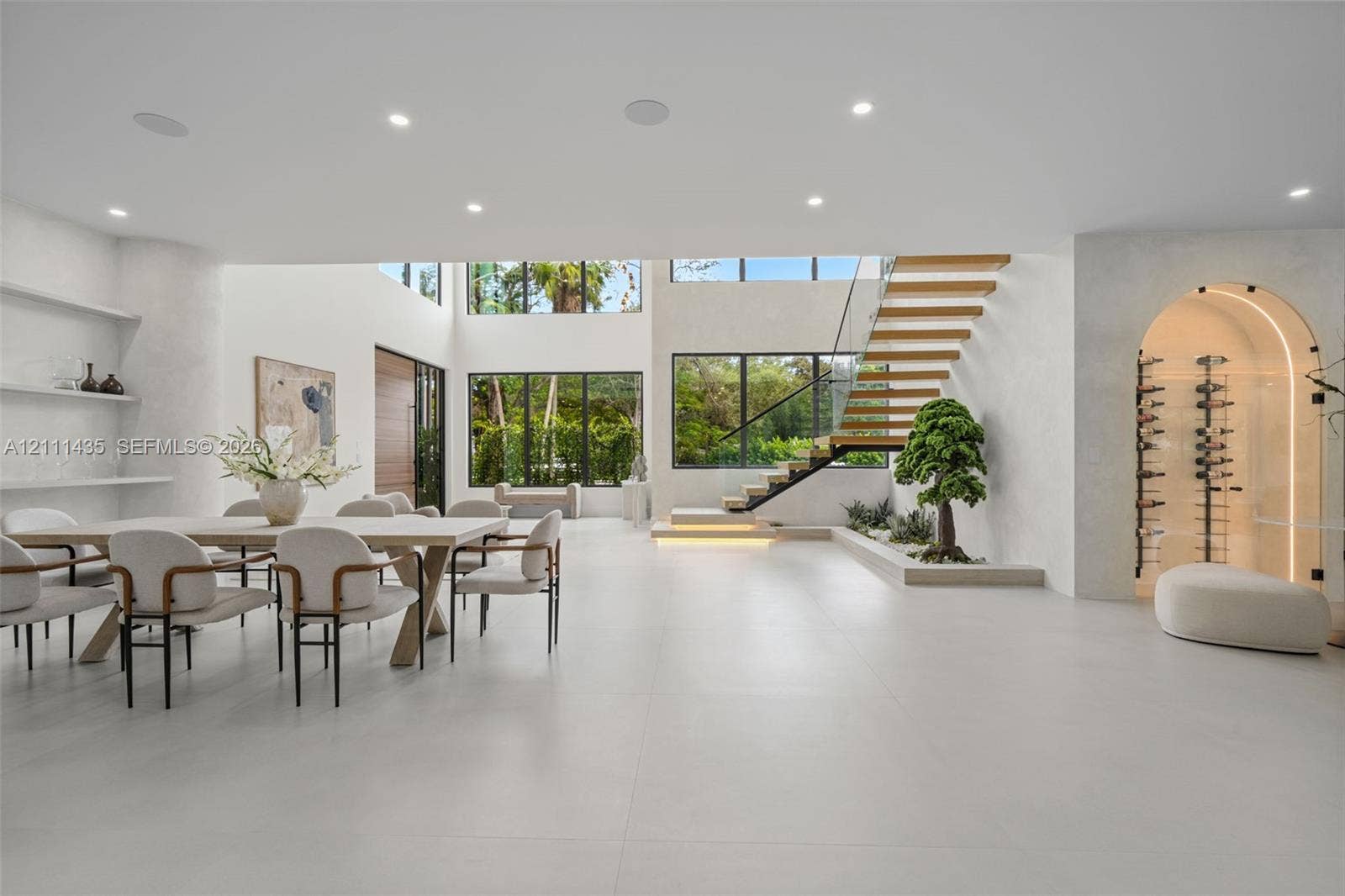 9530 SW 95th Court, Miami, FL 33176 Open-concept living space featuring large-format tile flooring, a floating staircase with glass railings, built-in recessed shelving, a glass-enclosed arched wine display, and expansive floor-to-ceiling windows