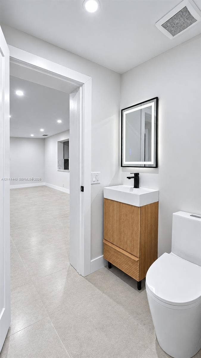 Modern vanity with wood-finish cabinetry, white basin, matte black faucet, and rectangular lighted mirror, complemented by ceramic tile flooring and a minimalist dual-flush toilet