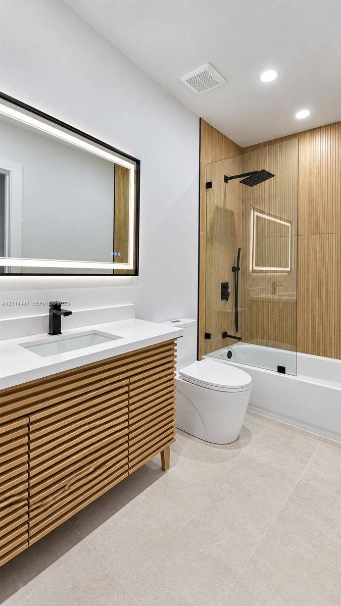 Slatted wood-finish vanity with a solid surface countertop, rectangular under-mount sink, matte black faucet, illuminated LED mirror, and tub-shower combination with wood-finish tile surround and frameless glass partition