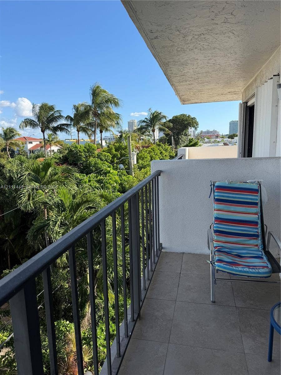 8255 Abbott Avenue, #402, Miami Beach, Miami Beach, FL 33141 Covered balcony with a dark metal railing, tiled flooring, and high-reaching exterior walls, complemented by mature palm trees and lush greenery