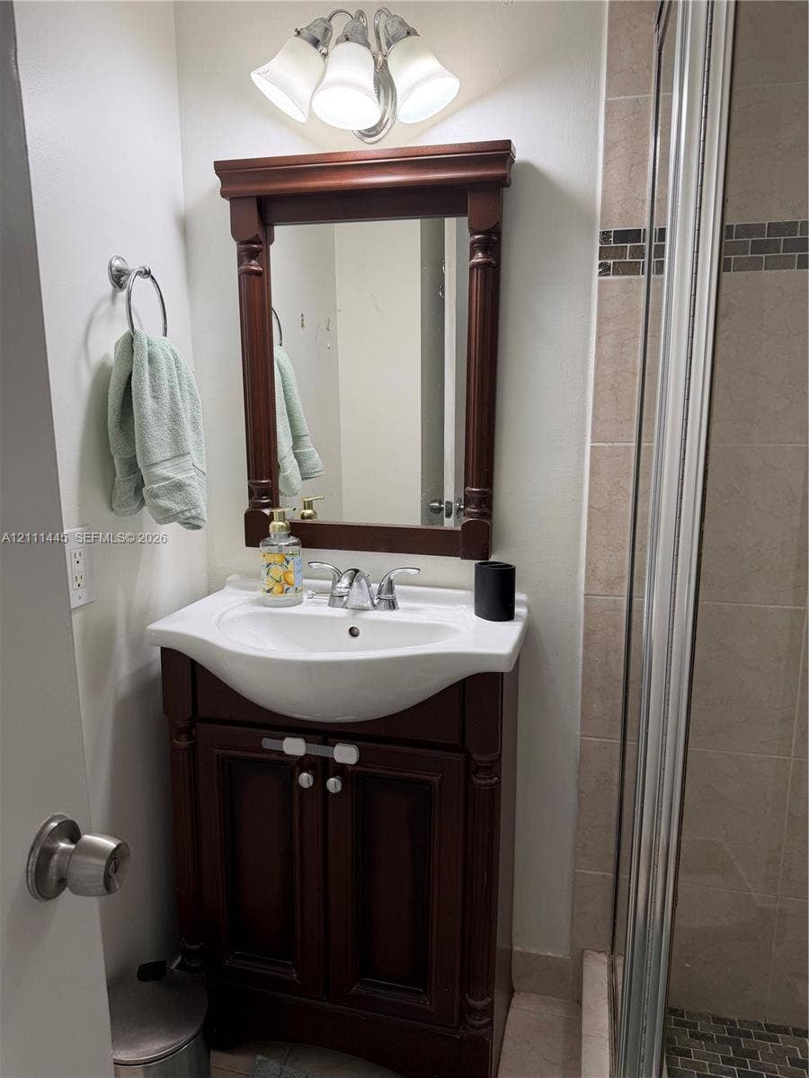8255 Abbott Avenue, #402, Miami Beach, Miami Beach, FL 33141 Wood-finish vanity with an integrated white sink, coordinated wood-frame mirror, three-light polished chrome fixture, and tiled shower stall with a decorative mosaic glass accent border