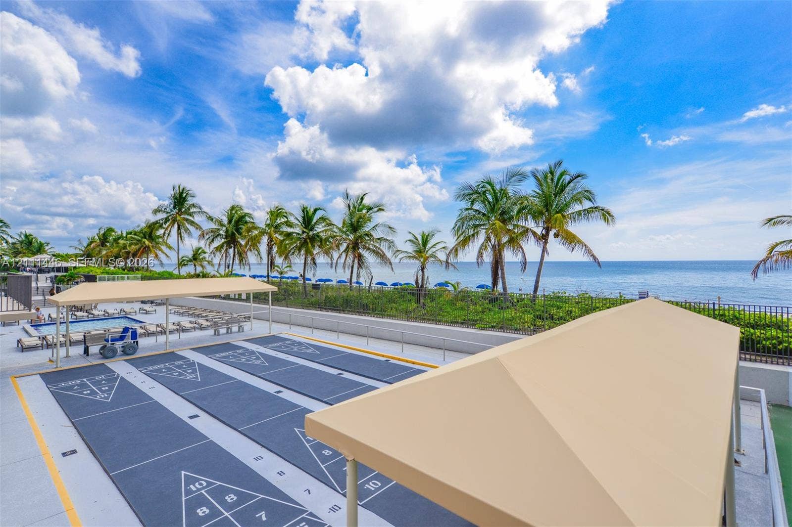 1965 S Ocean Drive, #15M, Hallandale Beach, Hallandale Beach, FL 33009 Recreational area featuring dual shuffleboard courts, covered seating structures, and perimeter fencing, set against a waterfront backdrop with mature palm trees