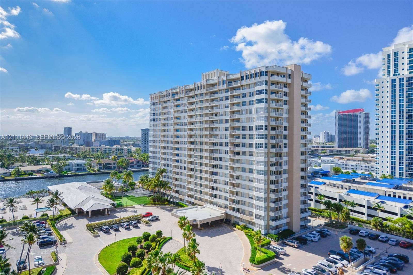 1965 S Ocean Drive, #15M, Hallandale Beach, Hallandale Beach, FL 33009 High-rise residential building featuring stacked private balconies, an expansive paved motor court with covered entrance, manicured landscaping, and water frontage