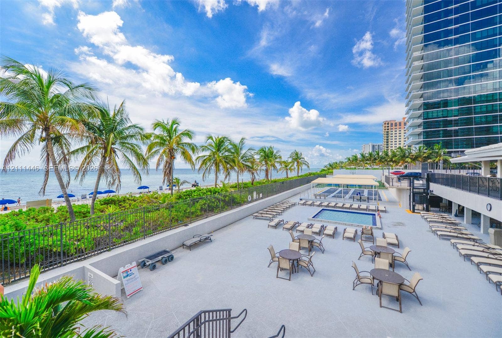 1965 S Ocean Drive, #15M, Hallandale Beach, Hallandale Beach, FL 33009 Expansive paved terrace featuring a swimming pool, shaded seating areas, metal perimeter fencing, and mature palm trees