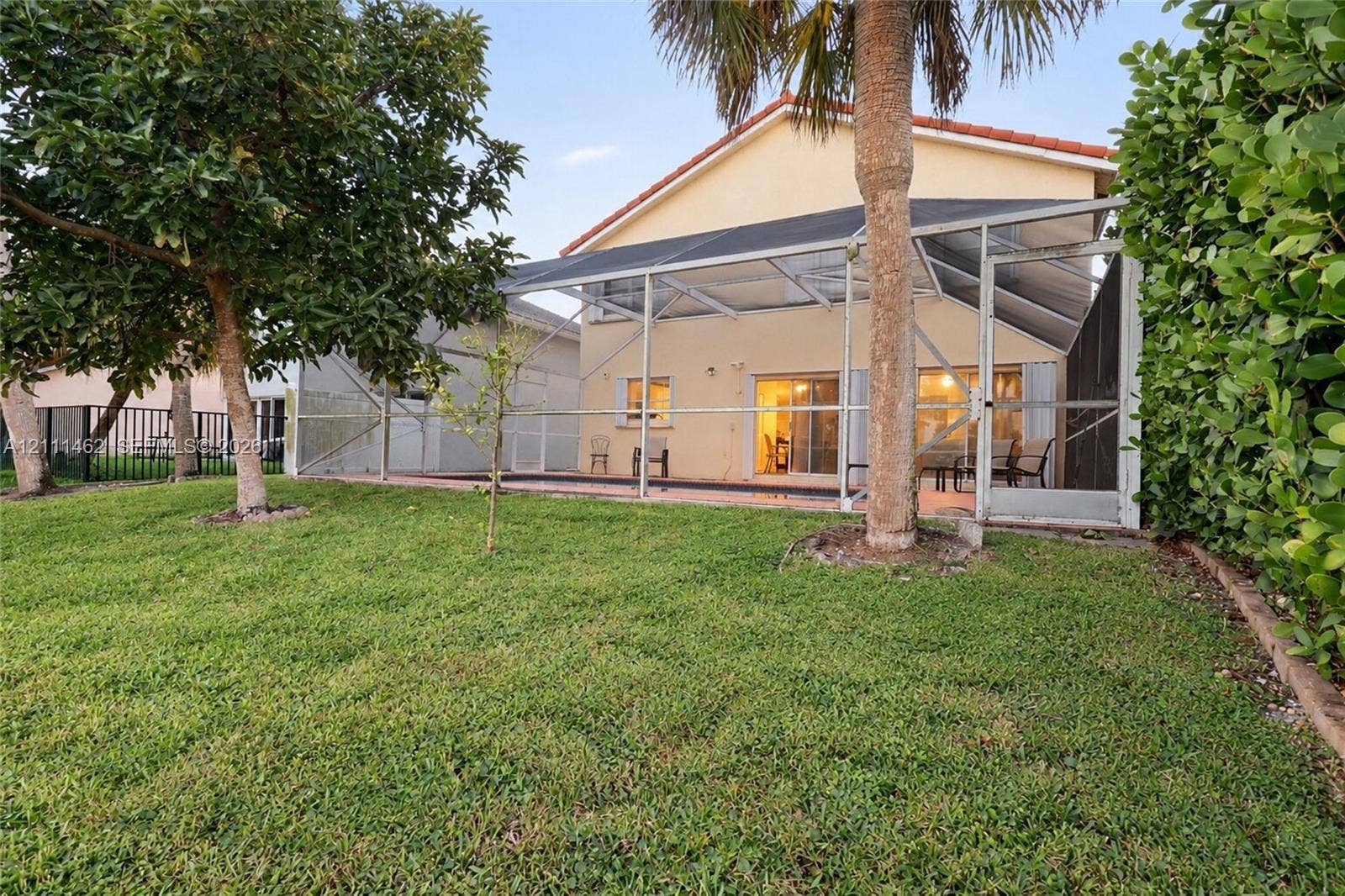 Rear exterior featuring a screened-in lanai, concrete patio, expansive lawn, mature trees, and privacy hedging