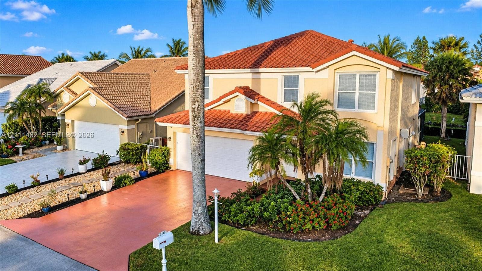 Two-story residence featuring a terracotta-tiled roof, stucco exterior, two-car garage, painted concrete driveway, and mature landscaping