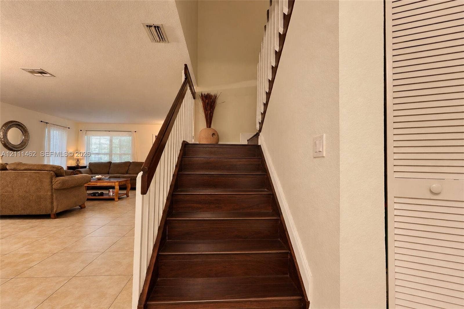 Staircase featuring dark wood-finish treads, white balusters, and a dark wood handrail, paired with neutral floor tiling and louvered closet doors