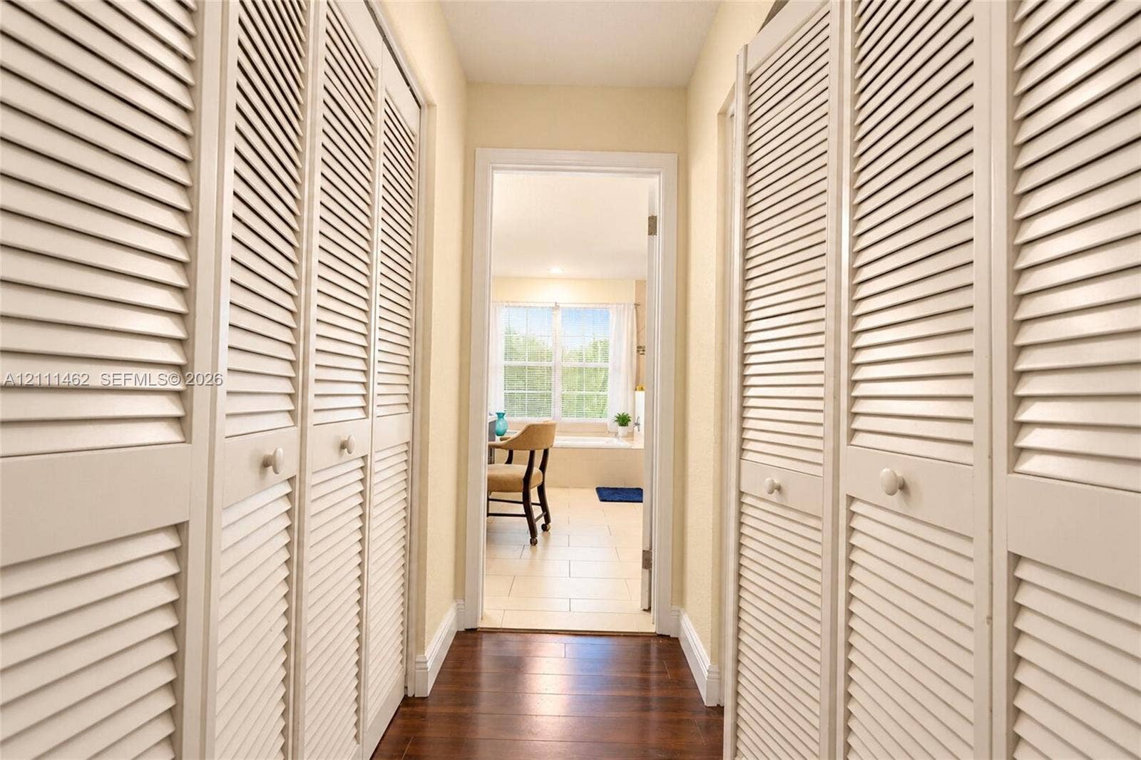 Hallway featuring full-height floor-to-ceiling louvered closet doors, wood-finish flooring, and a multi-pane window