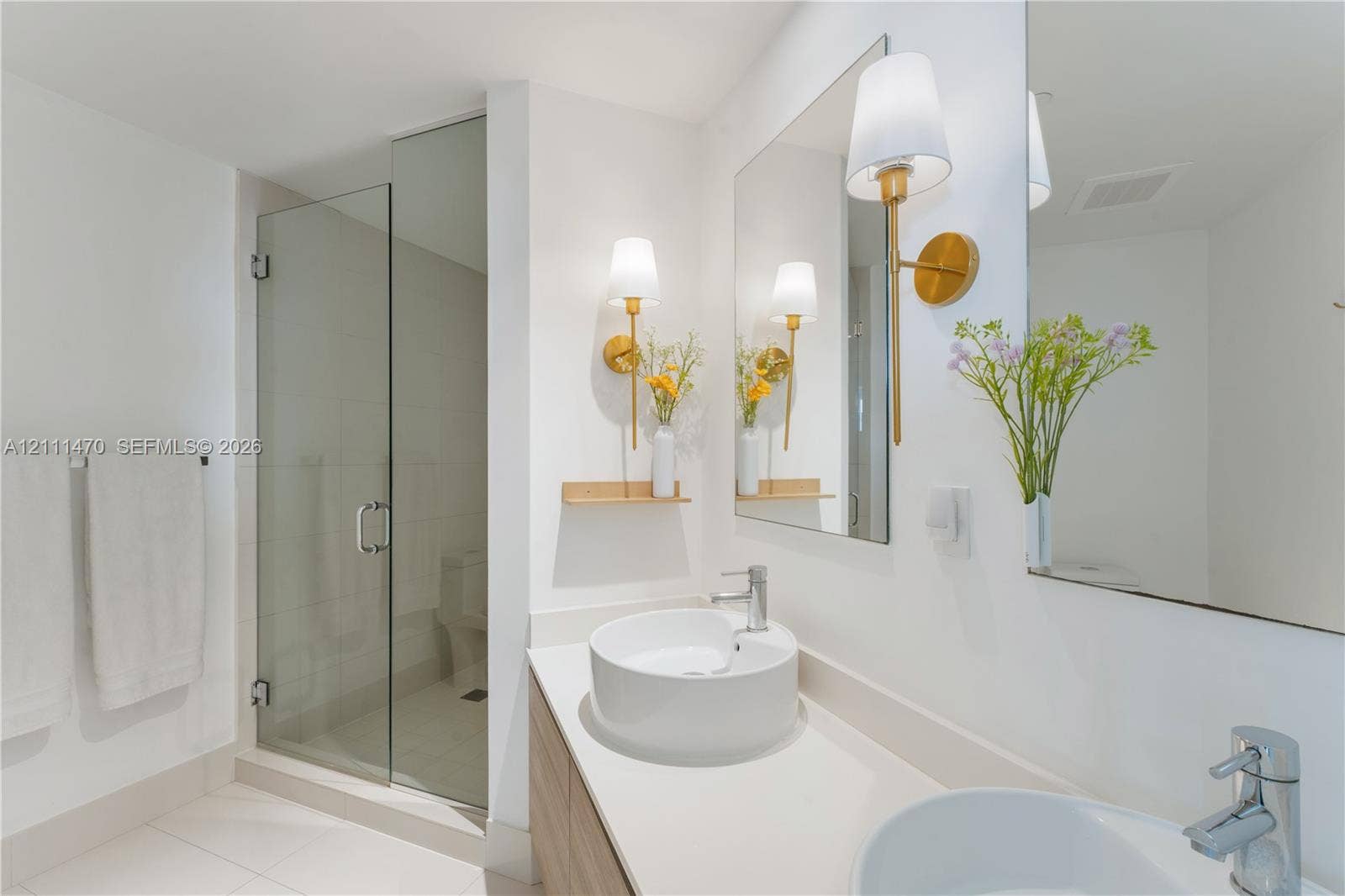 Bathroom featuring a dual-sink vanity with light-tone wood cabinetry, a quartz countertop, glass-enclosed shower, and gold-finish wall sconces