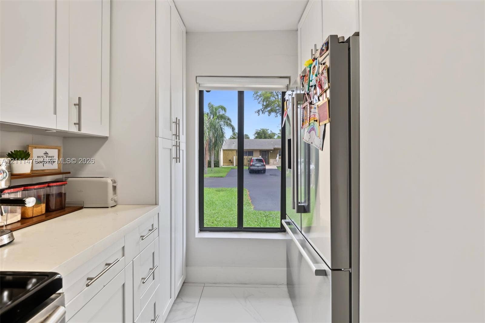 White shaker-style cabinetry, solid-surface countertops, floor-to-ceiling pantry storage, stainless steel refrigerator, and large-format tile flooring